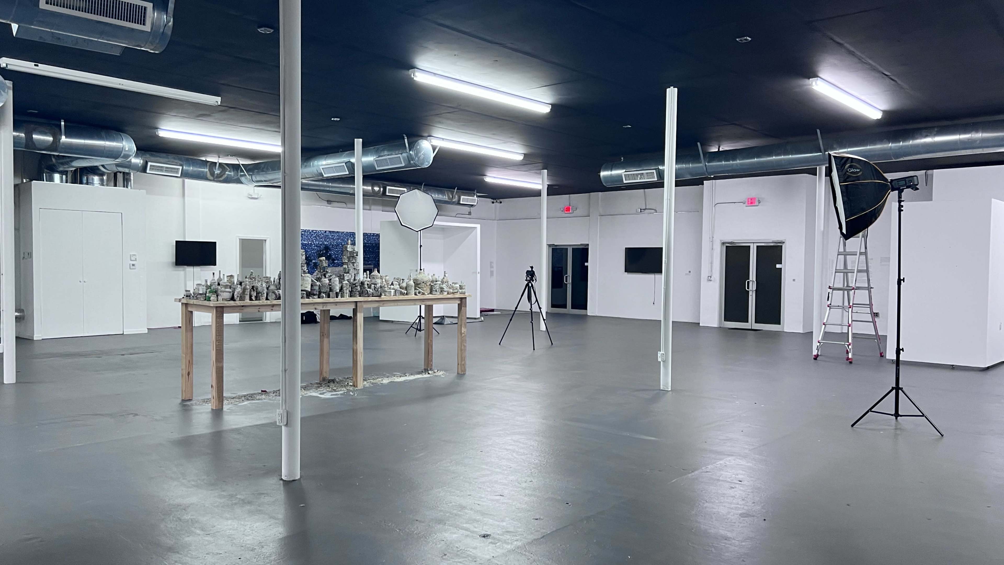 An empty industrial space features a central table with items arranged on it, photography equipment, and several light sources.