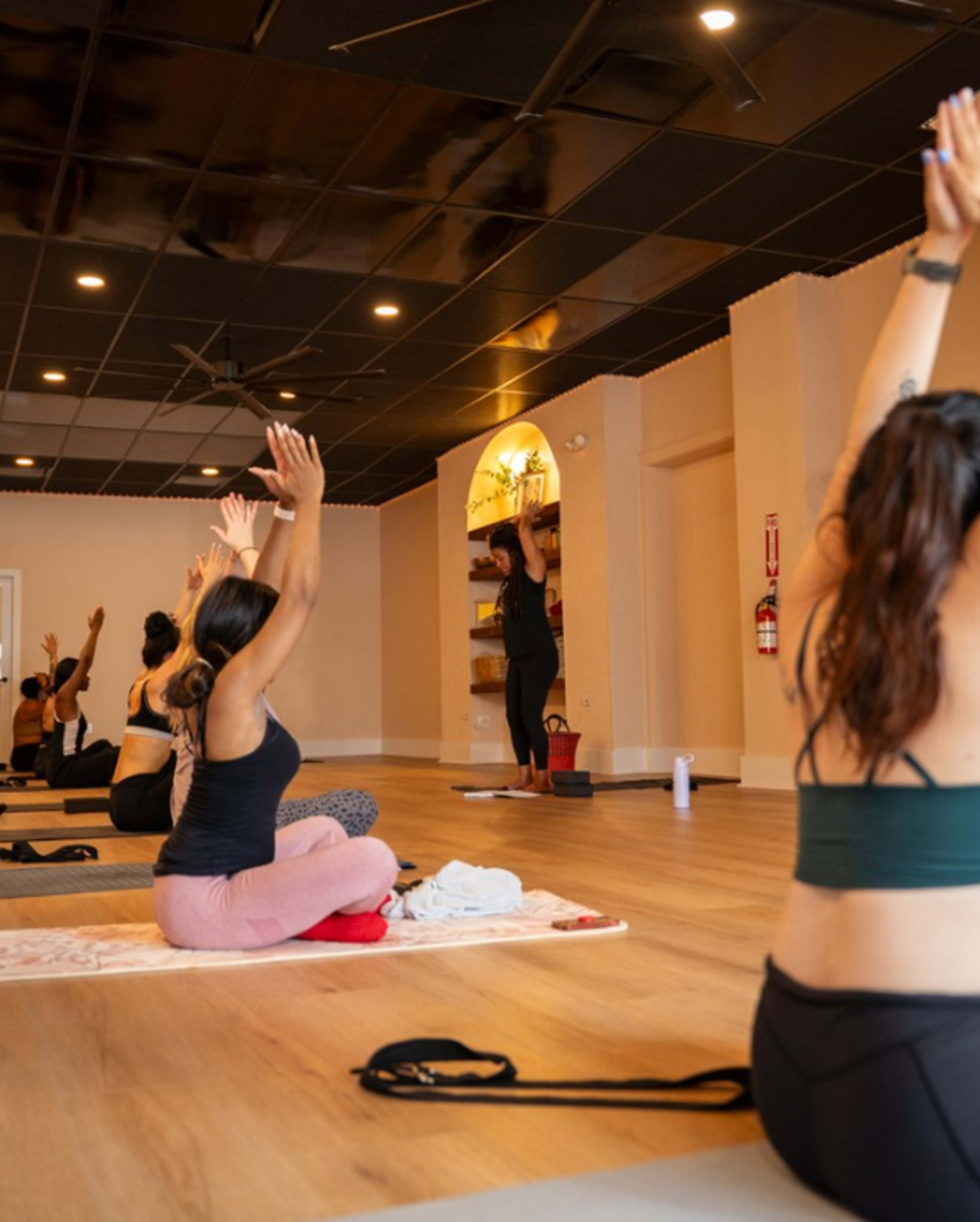 A group of individuals participates in a yoga class, seated on mats with arms raised, while an instructor demonstrates a pose in front of them.