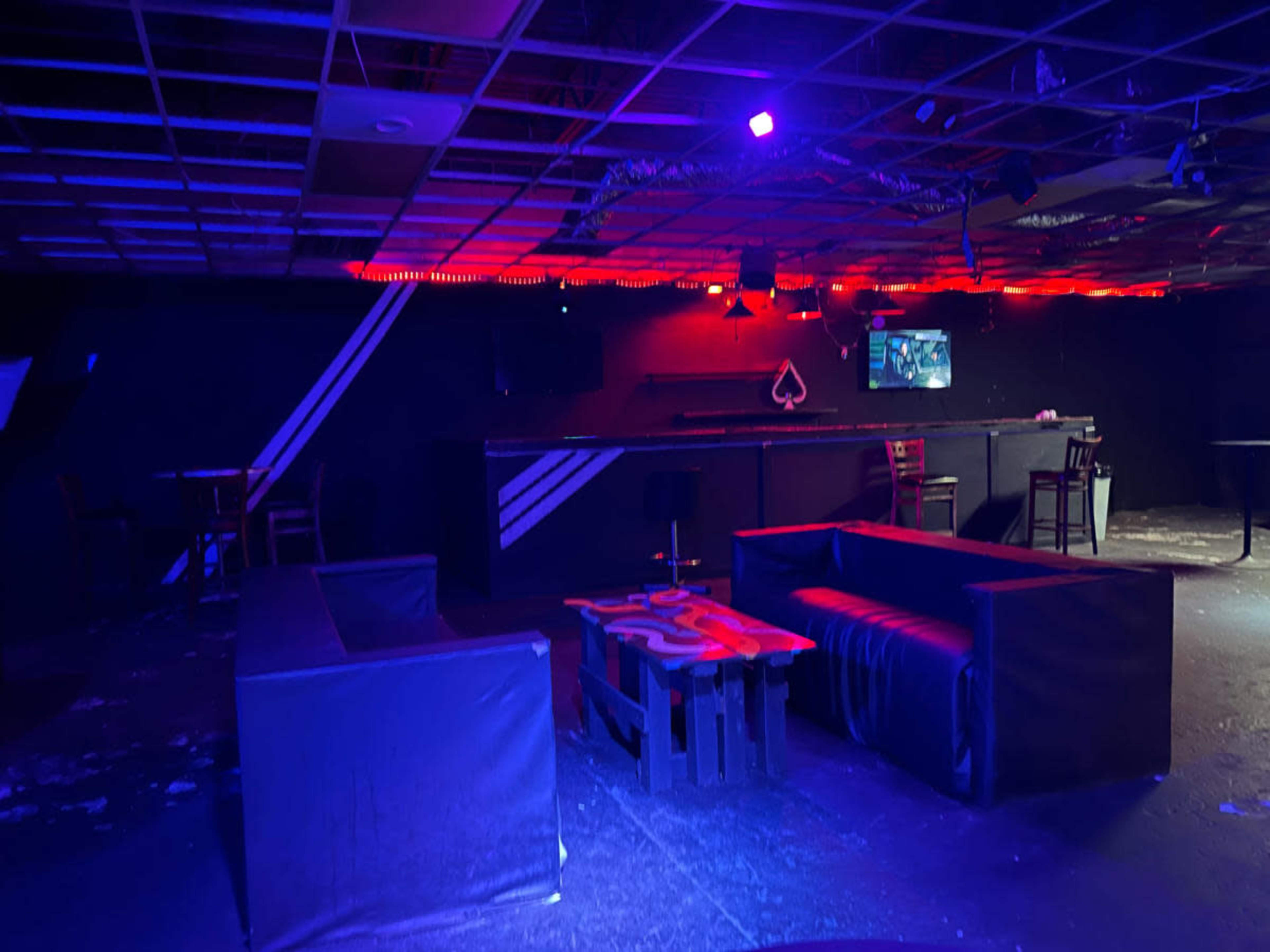The image shows an interior of a dimly lit lounge or nightclub featuring black furniture, a bar area with a TV, and purple and red lighting.