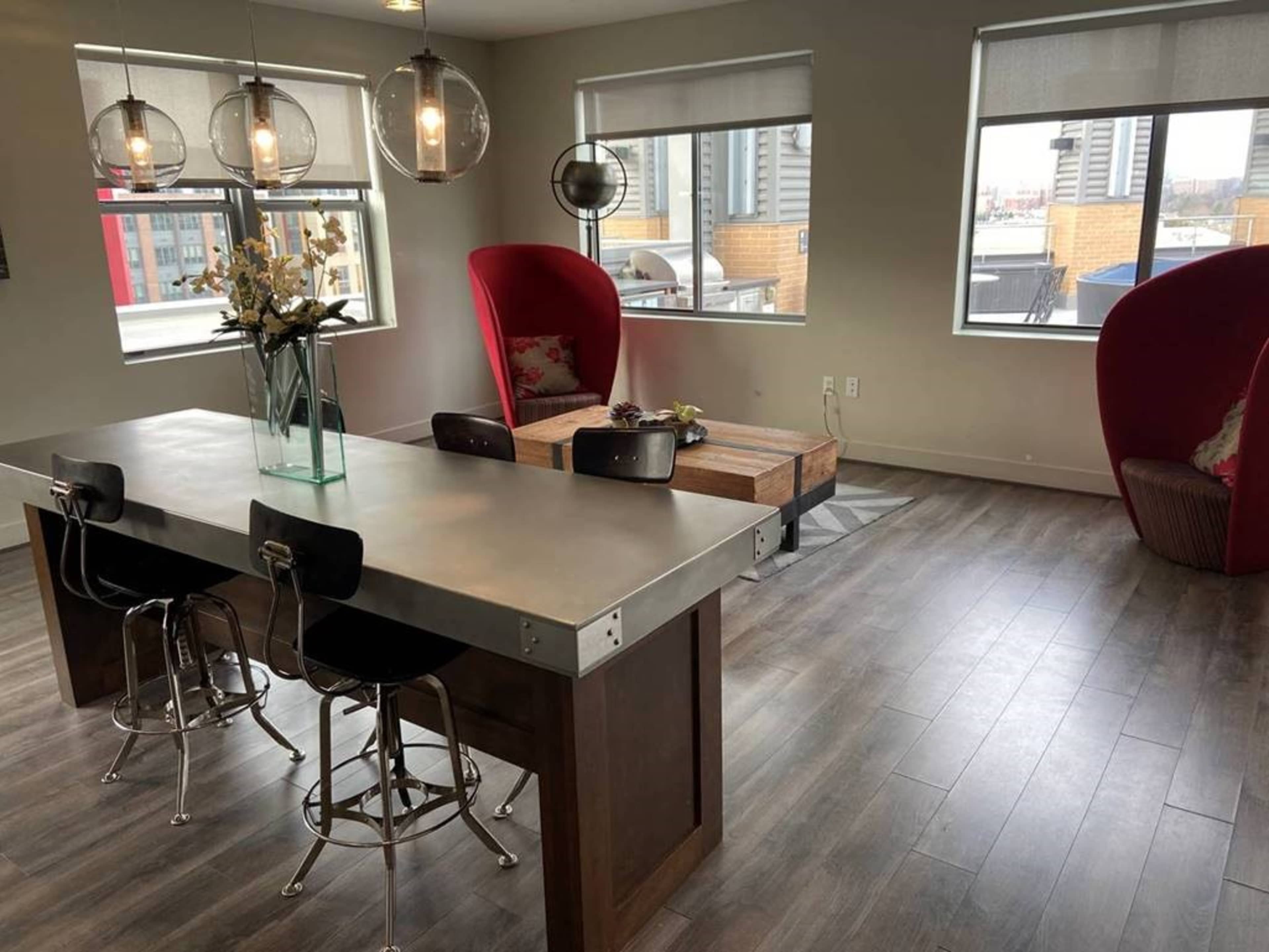 The image shows a modern interior featuring a dining area with a metal table and four stools, a wooden coffee table, and two large red chairs beside large windows.