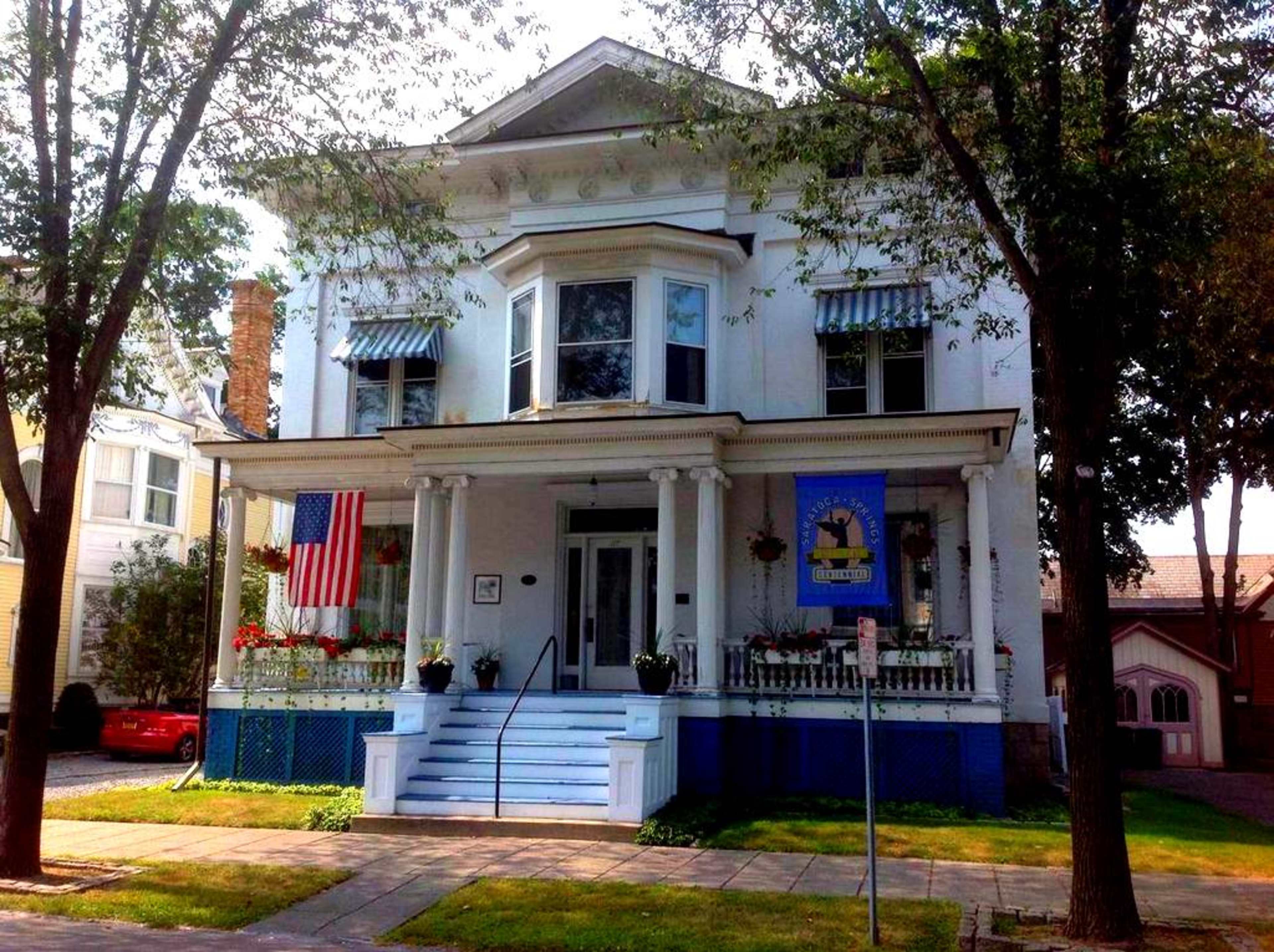 The image shows a large, two-story white house with a prominent porch, decorative elements, and an American flag hanging from the front.