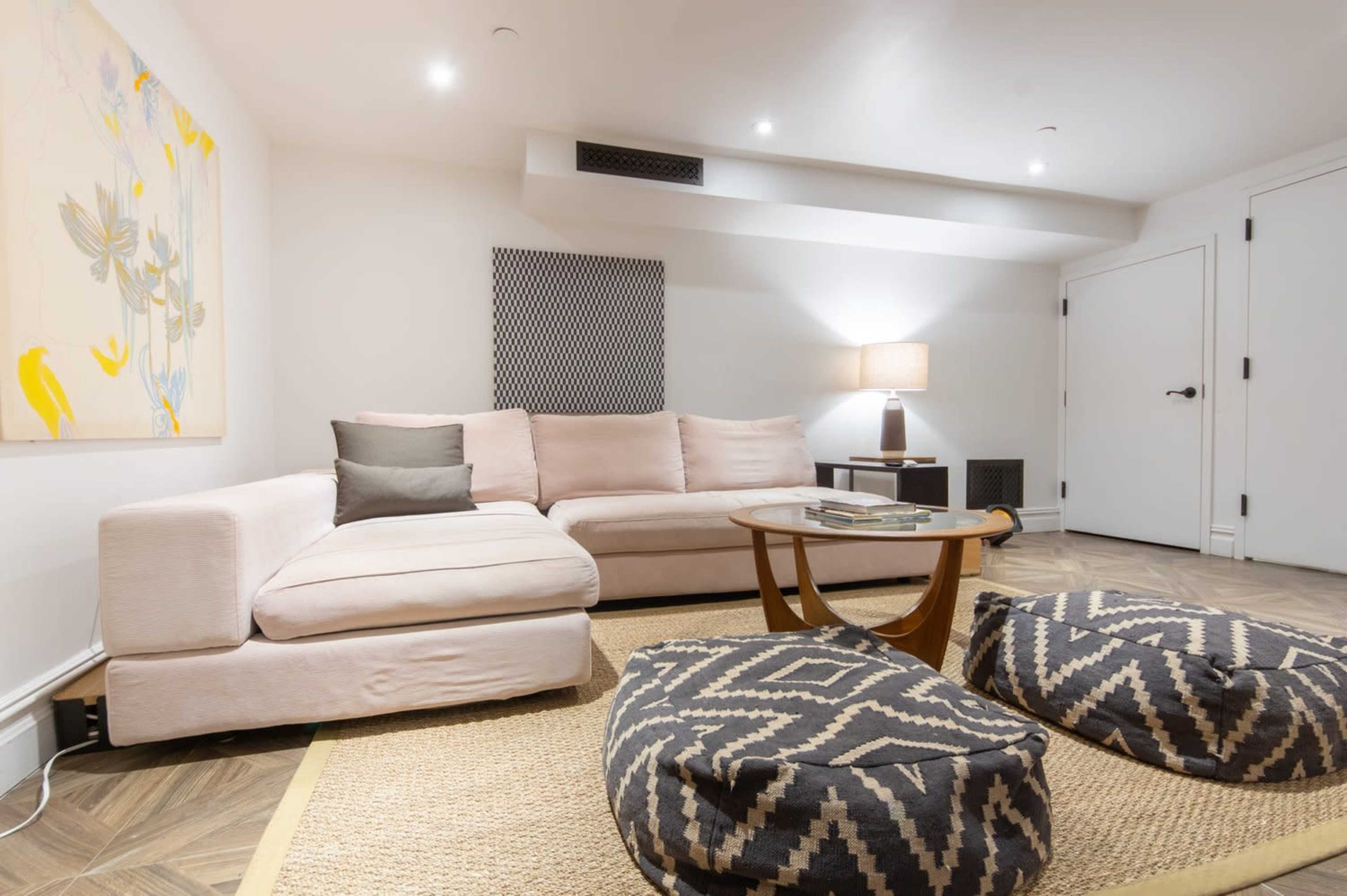 The image shows a modern living room featuring a light-colored sectional sofa, a round wooden coffee table, and two patterned poufs on a woven rug.