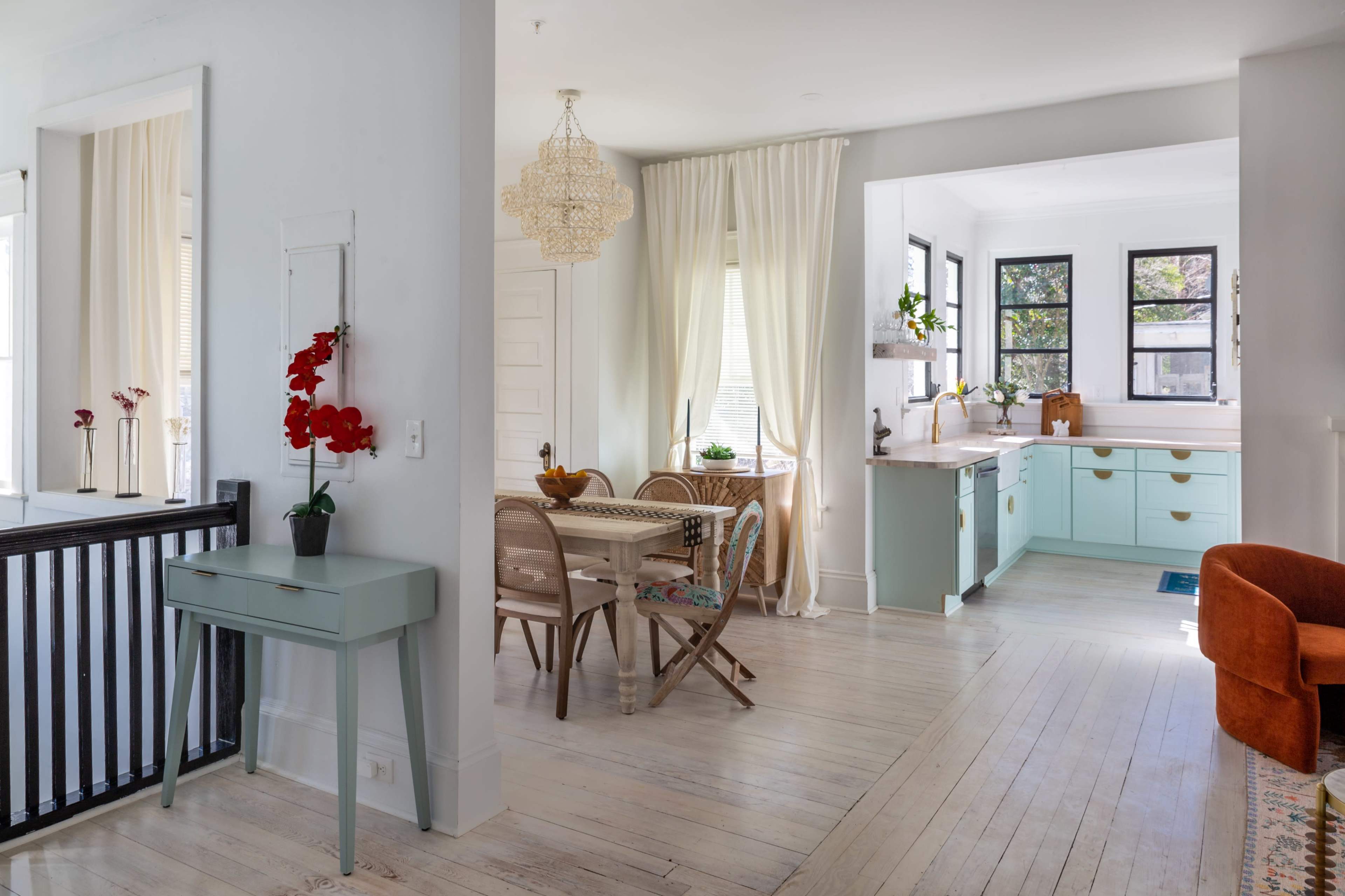 A brightly lit interior features a dining area with a wooden table and chairs, adjoining a modern kitchen with mint green cabinets and a large window.