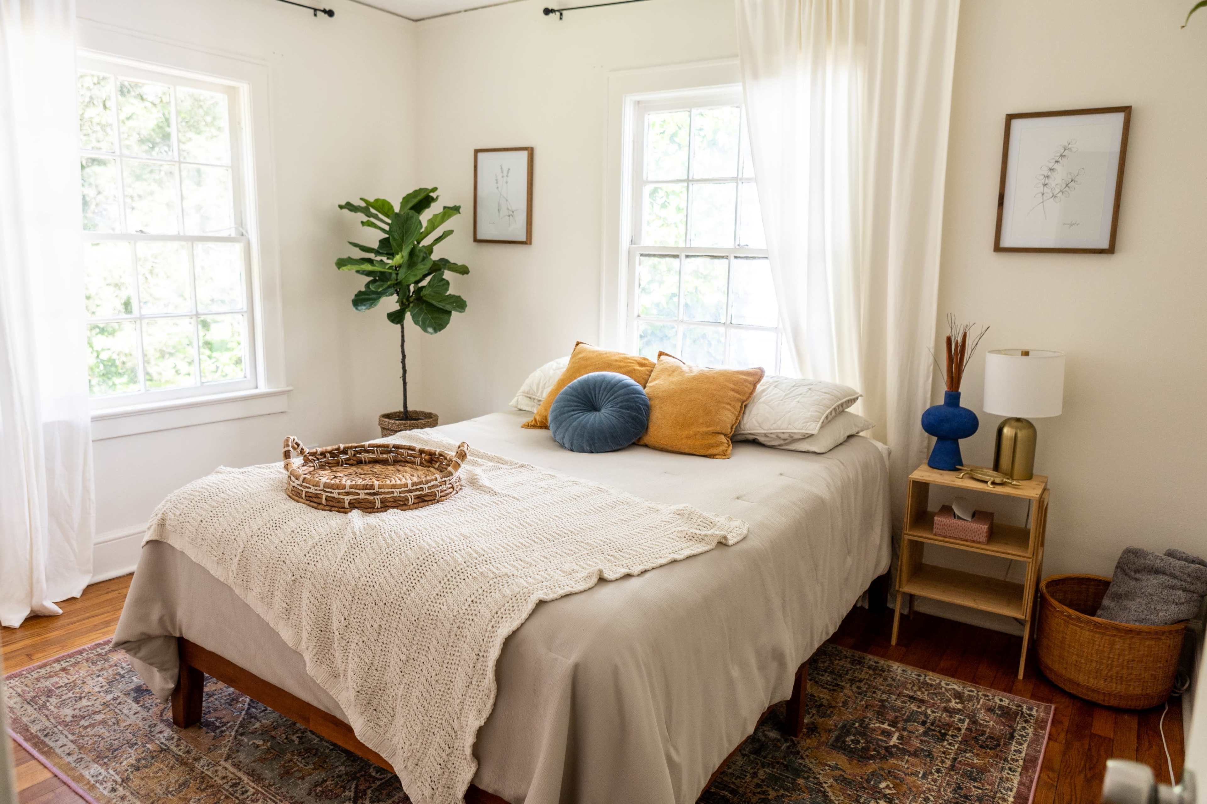 A cozy bedroom features a bed with decorative pillows, a textured throw, and a small bedside table beside large windows.