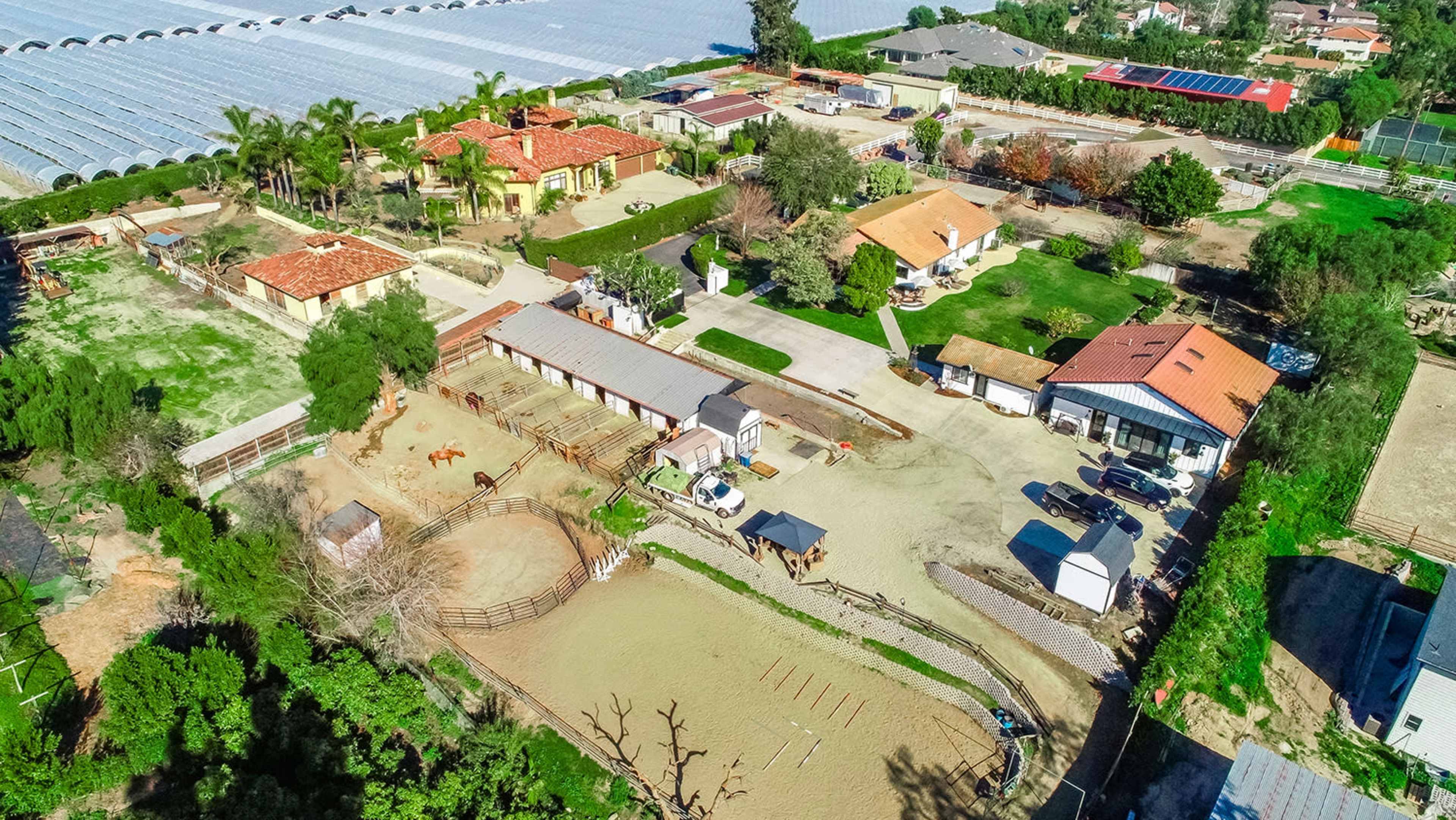 This aerial image shows a rural property with several buildings, horse stables, and a riding arena surrounded by greenery and a residential area.