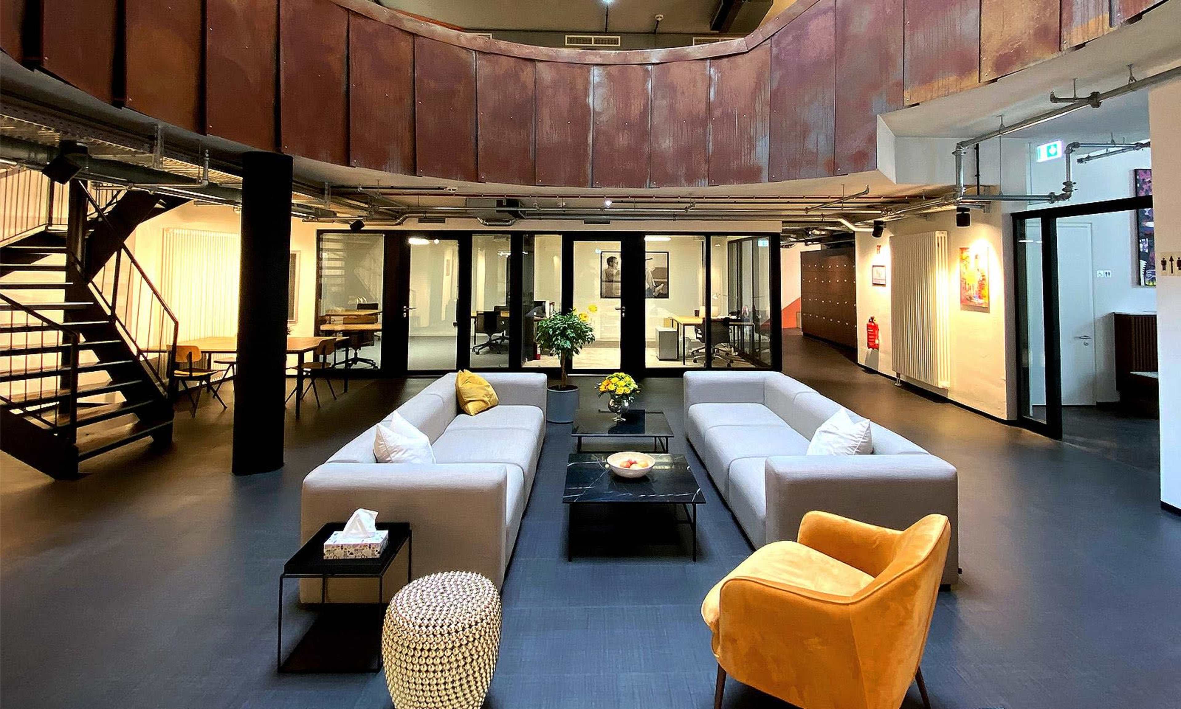 The image shows a modern open-plan lounge area with two gray sofas facing each other, a glass coffee table in the center, a yellow accent chair, and a staircase leading to an upper level.