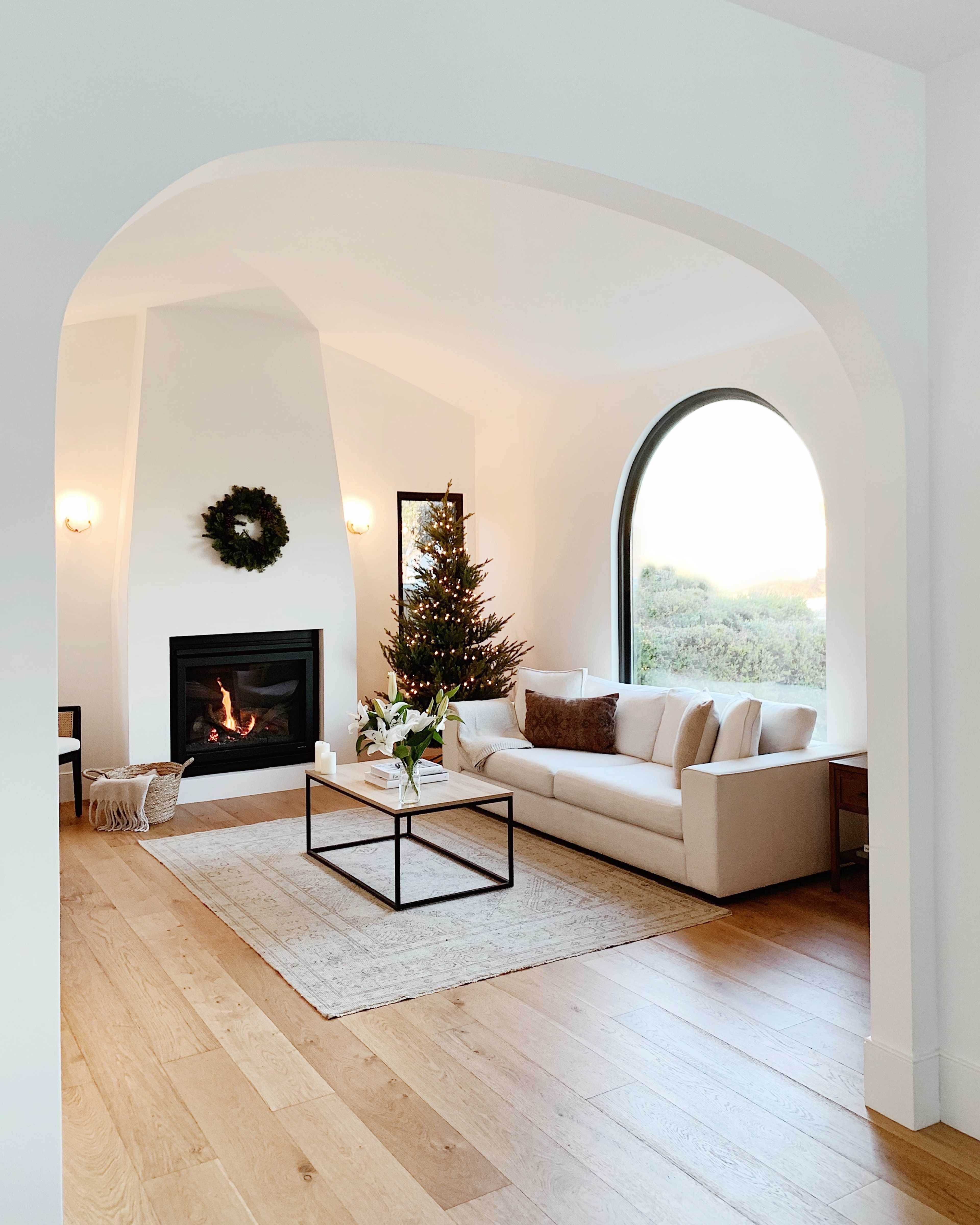 A modern living room features a white sofa, a coffee table, a Christmas tree, and a fireplace, with an archway leading to a window that overlooks greenery.