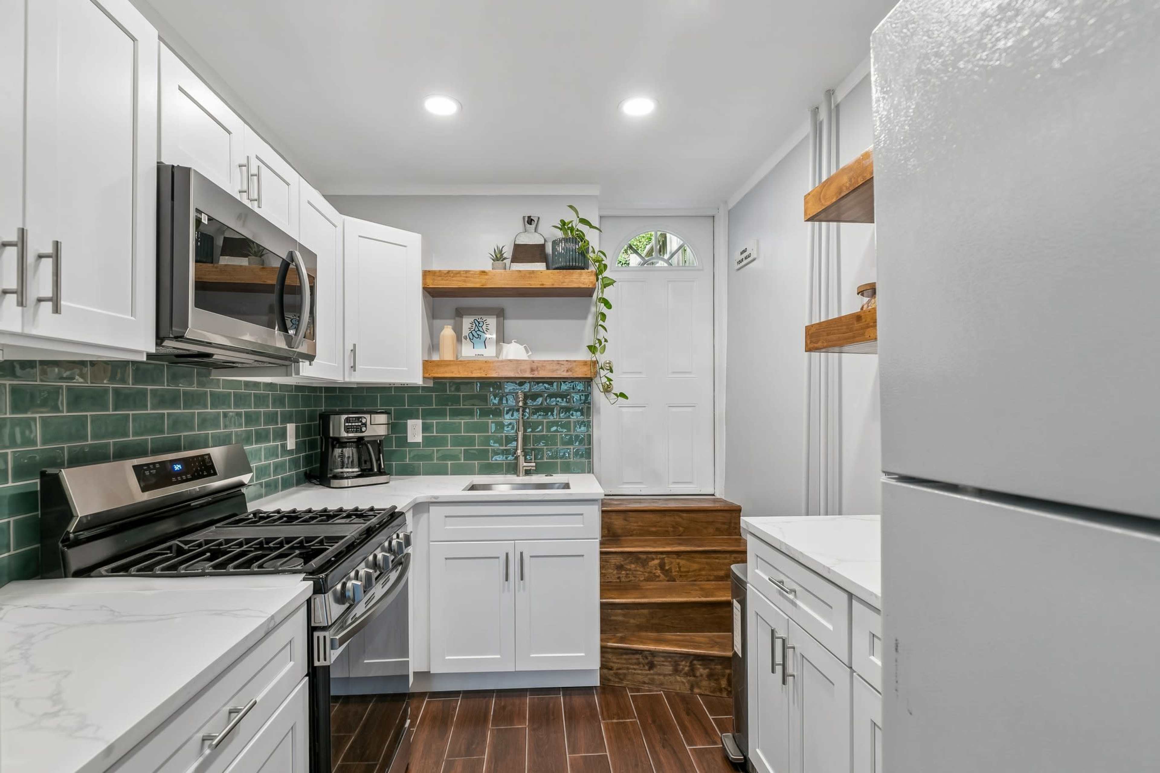 The image shows a modern kitchen featuring white cabinetry, a stainless steel oven and microwave, green tile backsplash, and open shelves with decorative items.