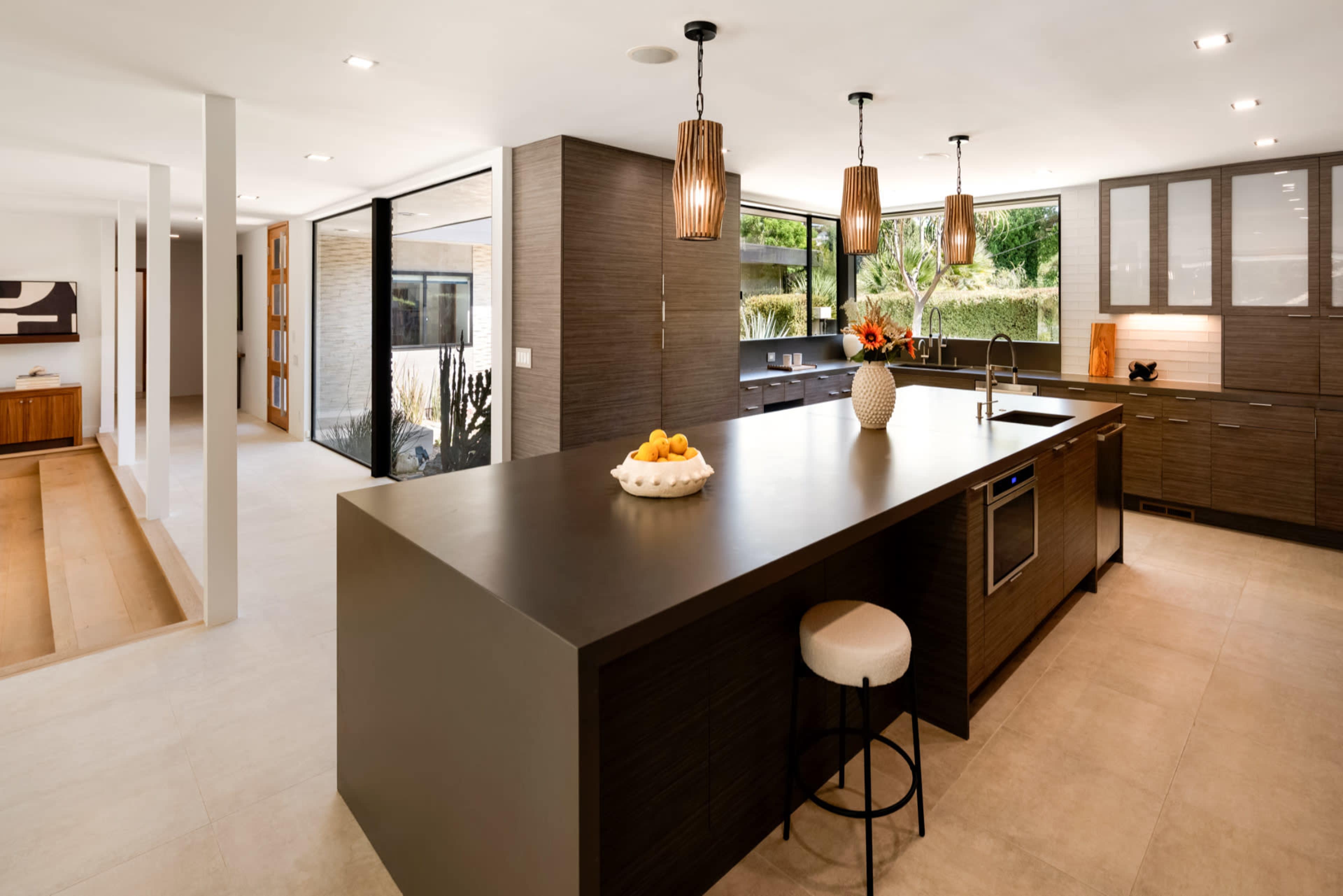 The image shows a modern kitchen with an island, dark cabinetry, and pendant lighting, connected to a bright living area with large windows.
