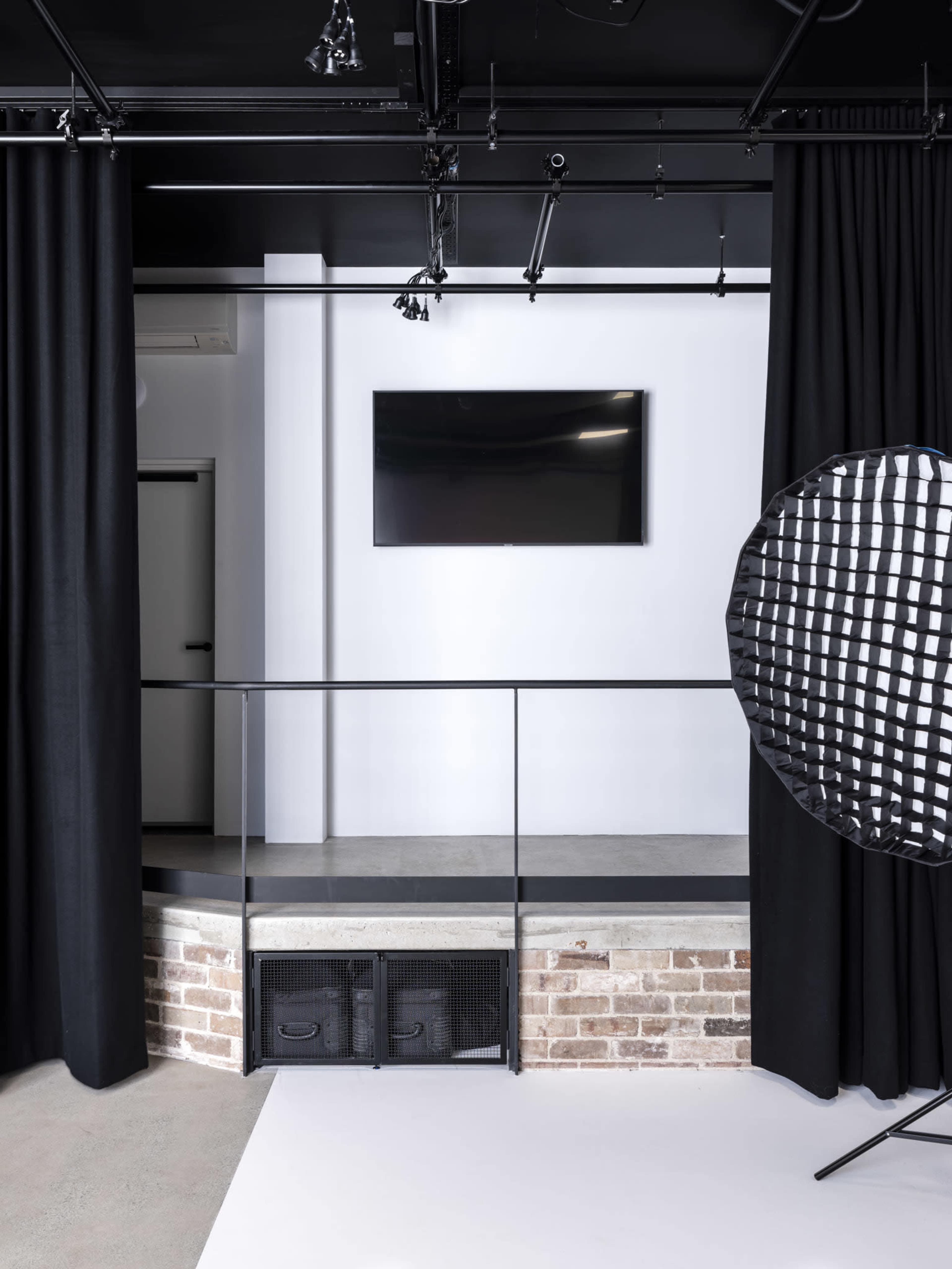 The image features a minimalist studio space with black curtains, a mounted television on the white wall, and a spotlight setup near a brick platform.
