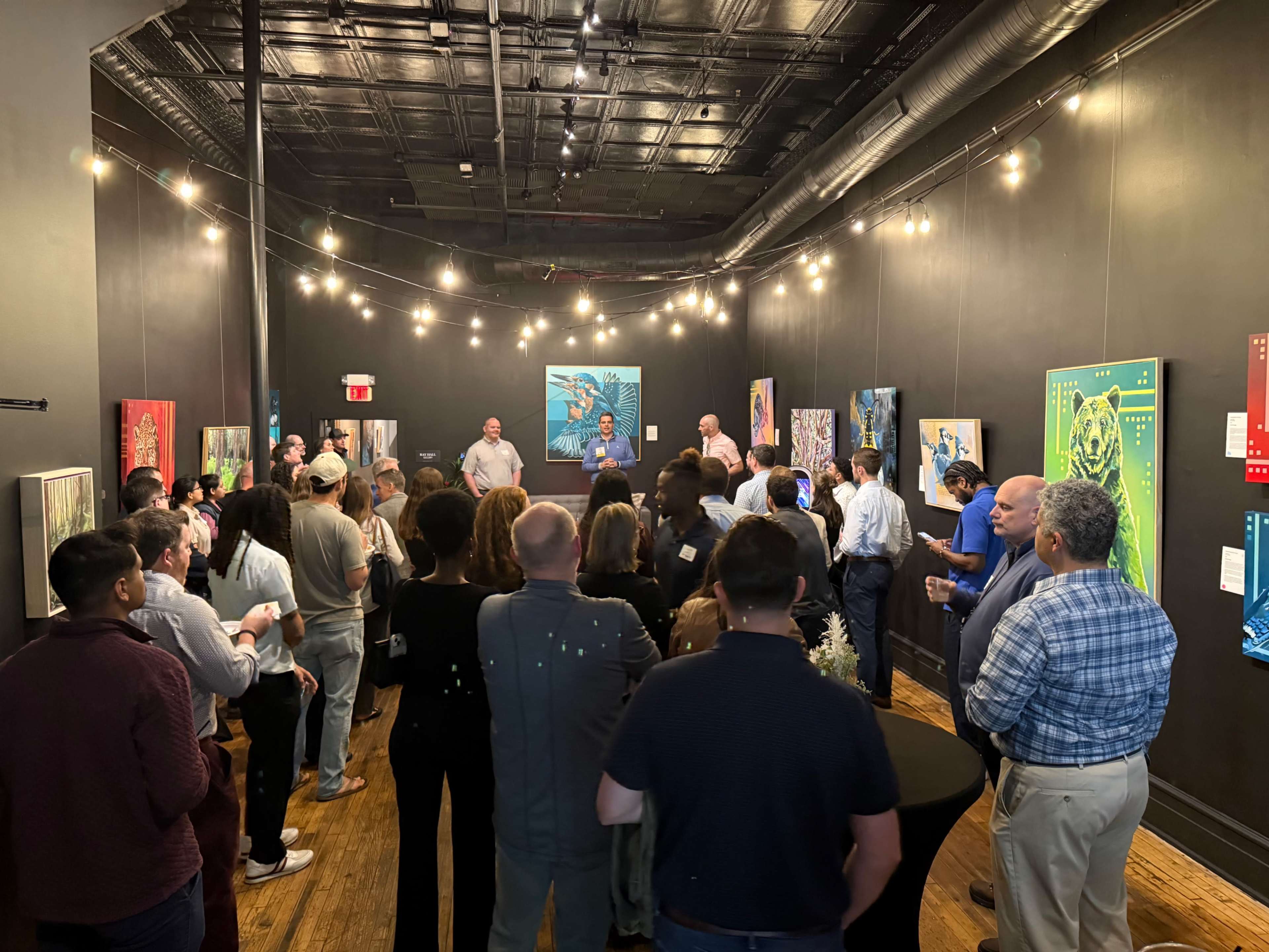 A crowd gathers in a gallery space adorned with colorful artwork, while a speaker addresses the audience under string lights.