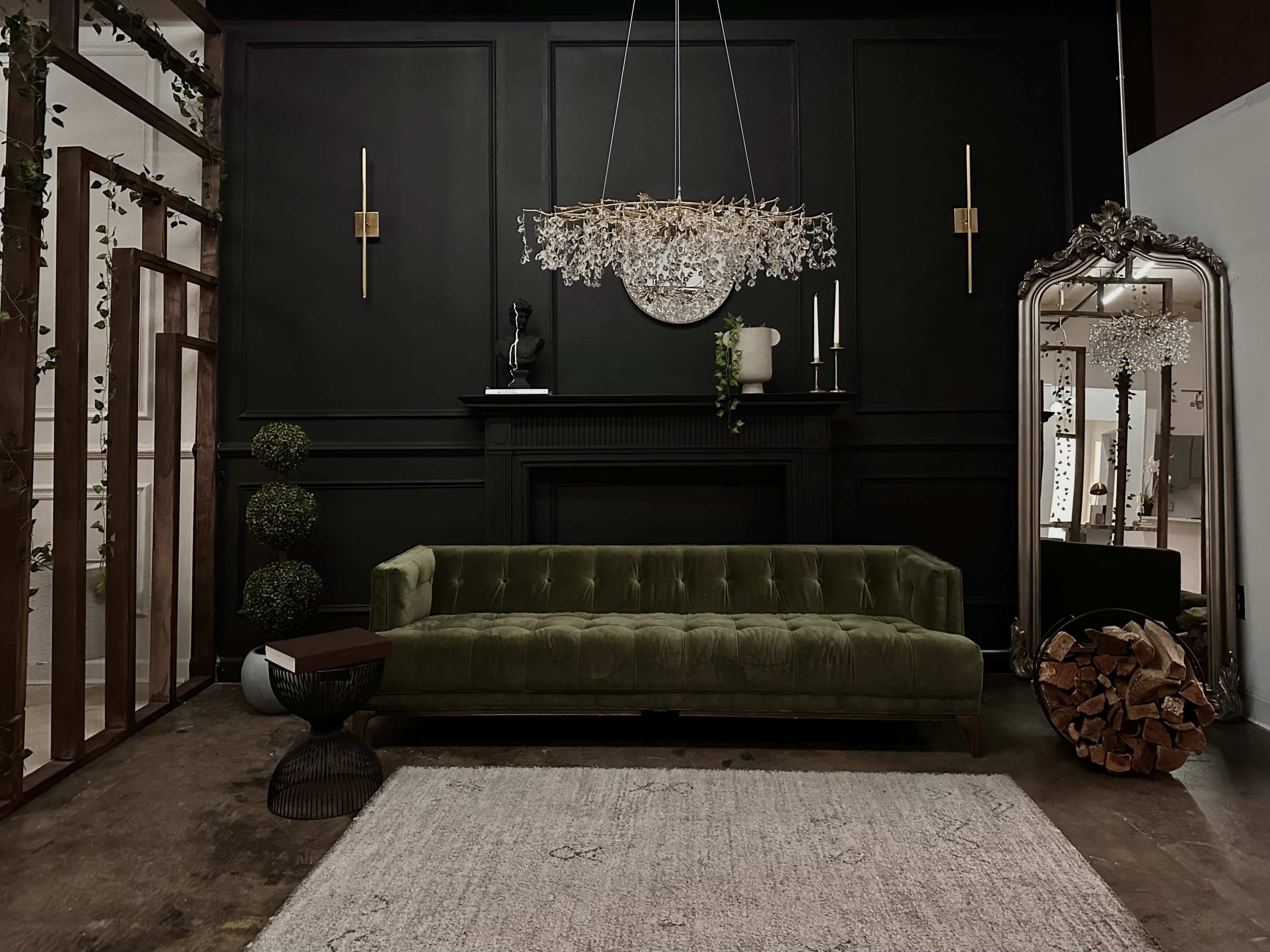 A green velvet sofa in front of a black wall, accompanied by a chandelier, decorative plants, a large mirror, and a log holder.