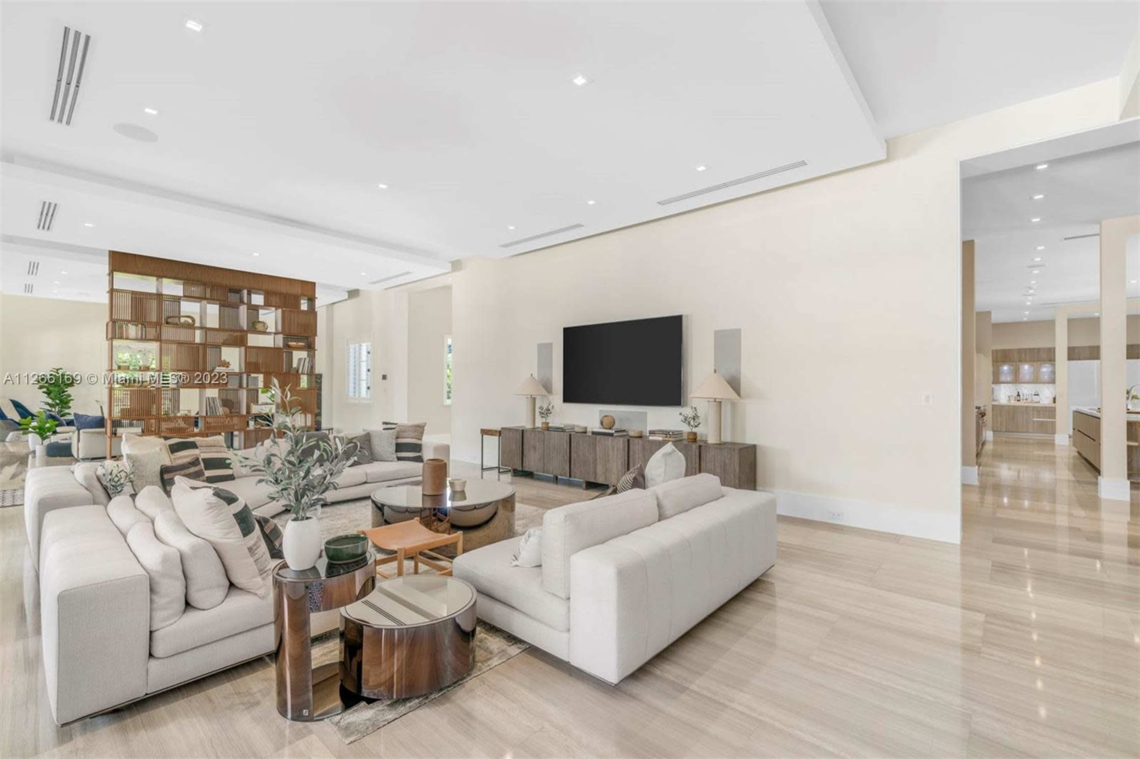 A spacious living room with light-colored walls, modern furniture, and a large television mounted on the wall, complemented by an open kitchen in the background.