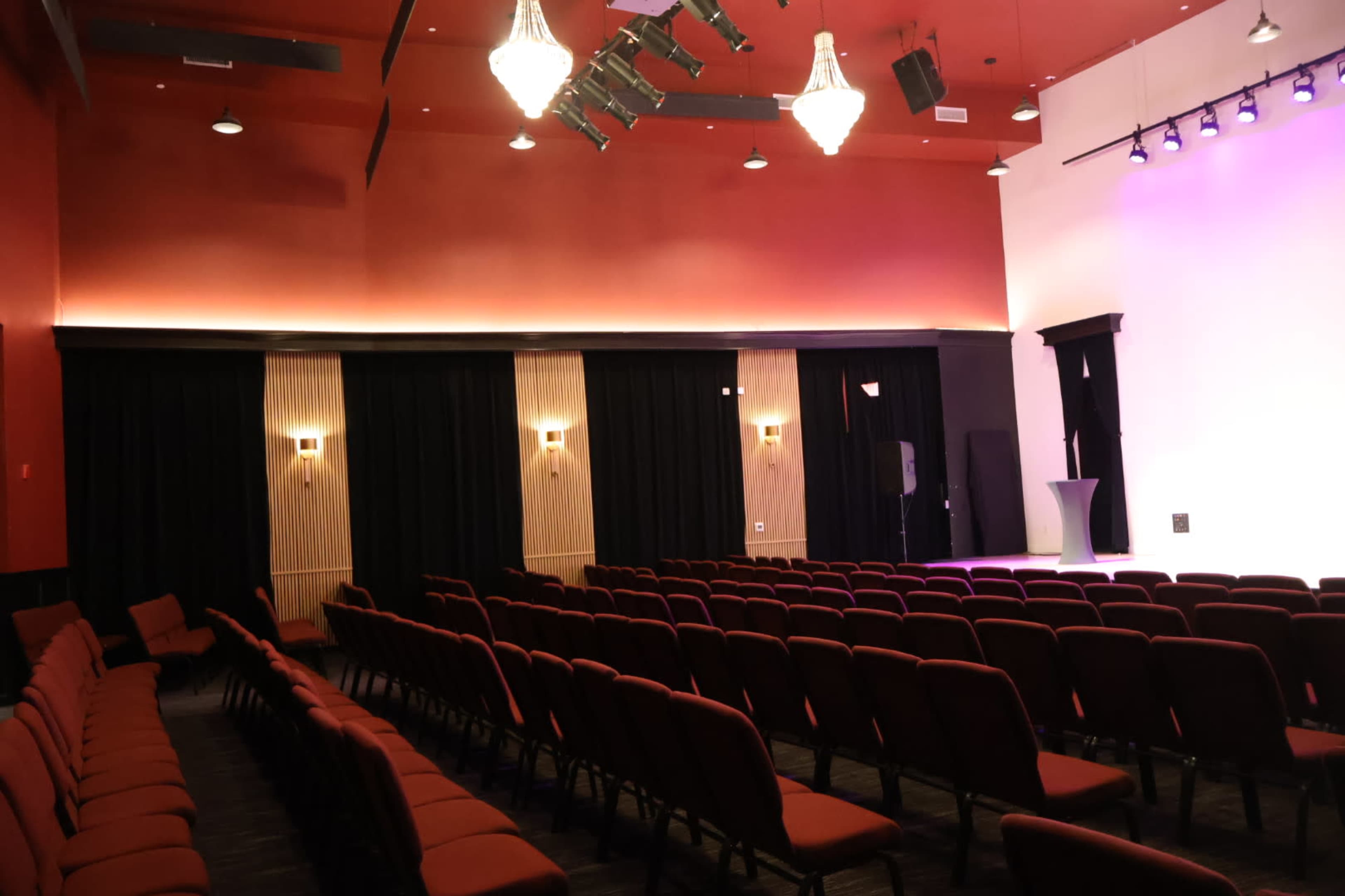 The image shows a theater interior with red seating arranged in rows facing a stage, illuminated by stage lights and featuring black curtains on the sides.