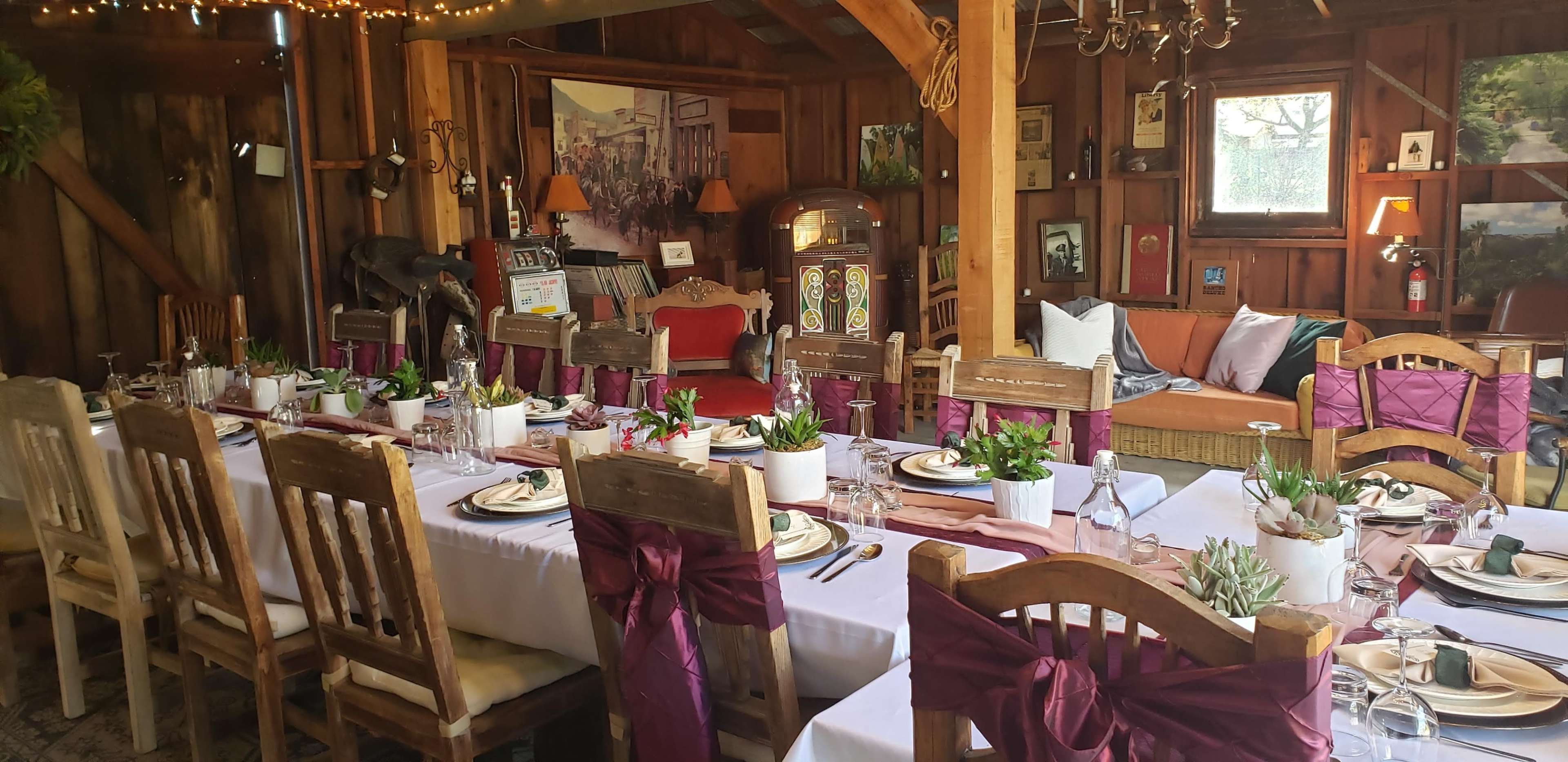 A long dining table is set with plates and glassware, surrounded by wooden chairs adorned with burgundy sashes, in a rustic indoor space featuring wooden beams and vintage decor.