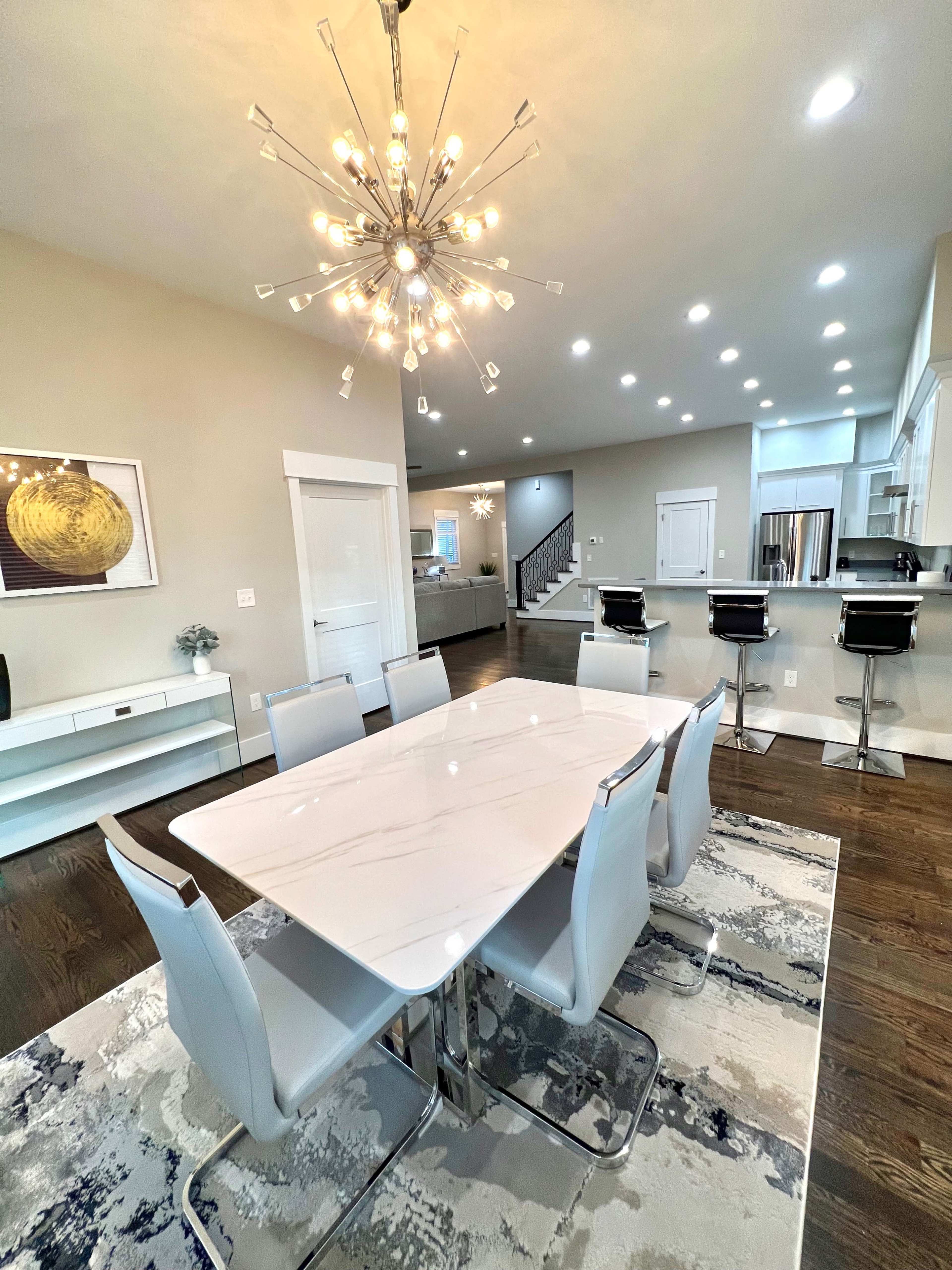A modern dining area features a marble table surrounded by six light-colored chairs, with a large chandelier overhead and an open kitchen visible in the background.