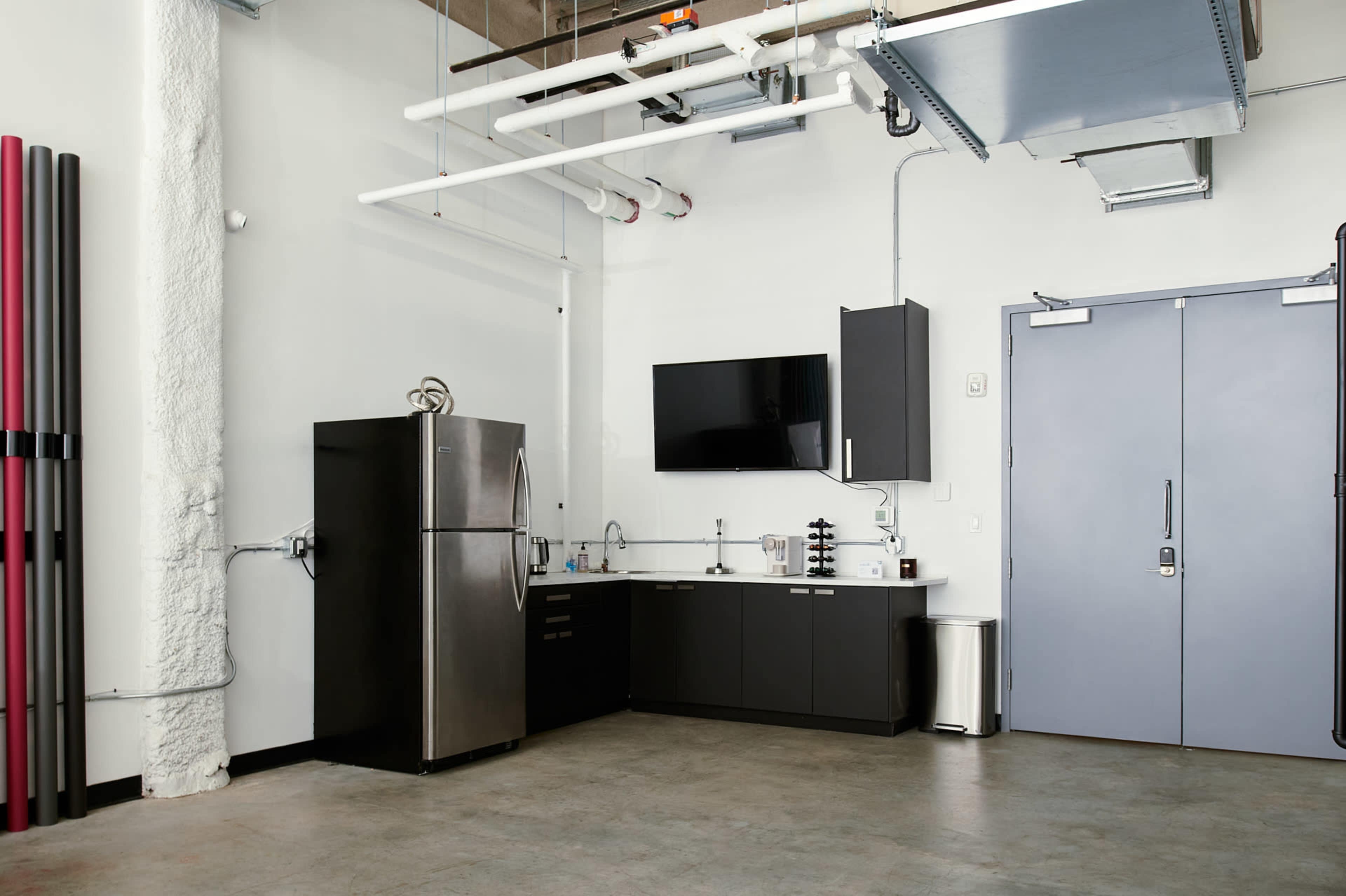 The image shows a kitchen space with a stainless steel refrigerator, dark cabinetry, a wall-mounted television, and a gray door in a modern industrial setting.