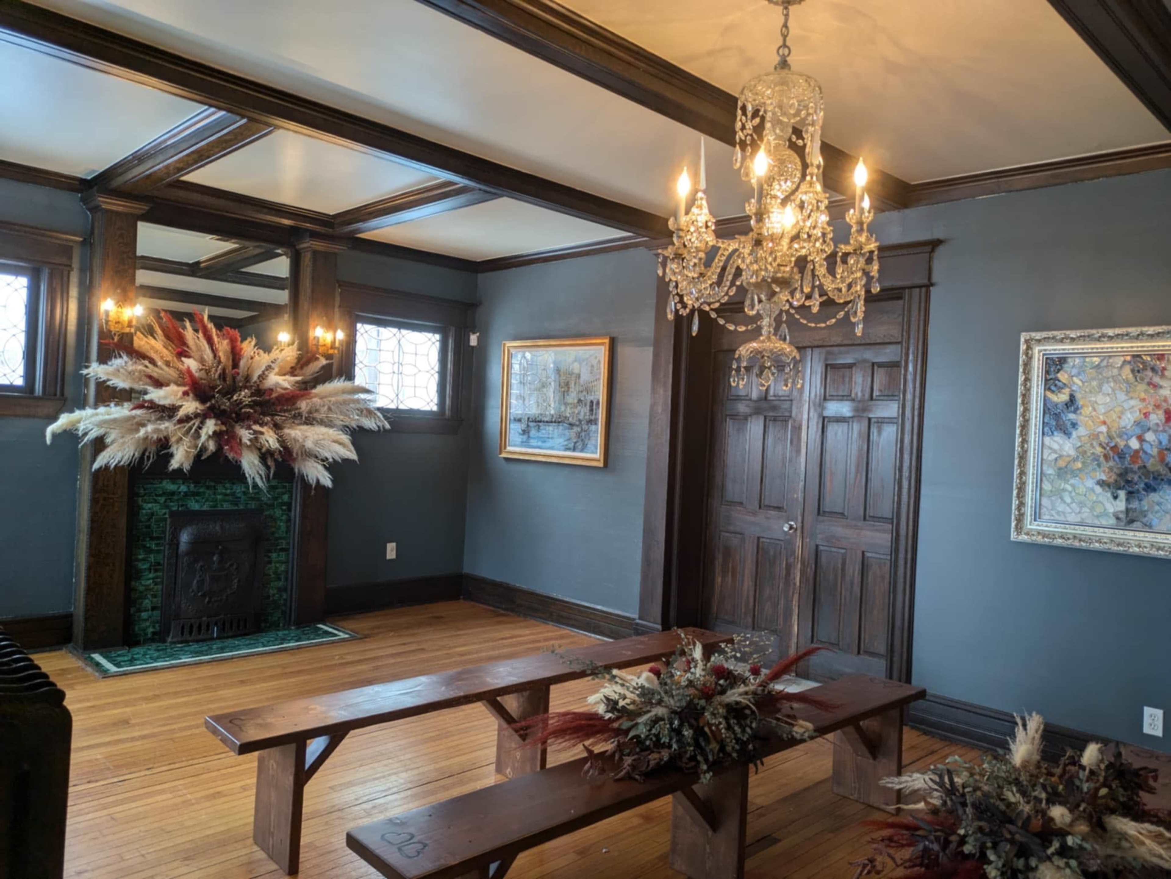 The room features wooden floors, paneled walls, a chandelier, and a decorative fireplace adorned with floral arrangements.