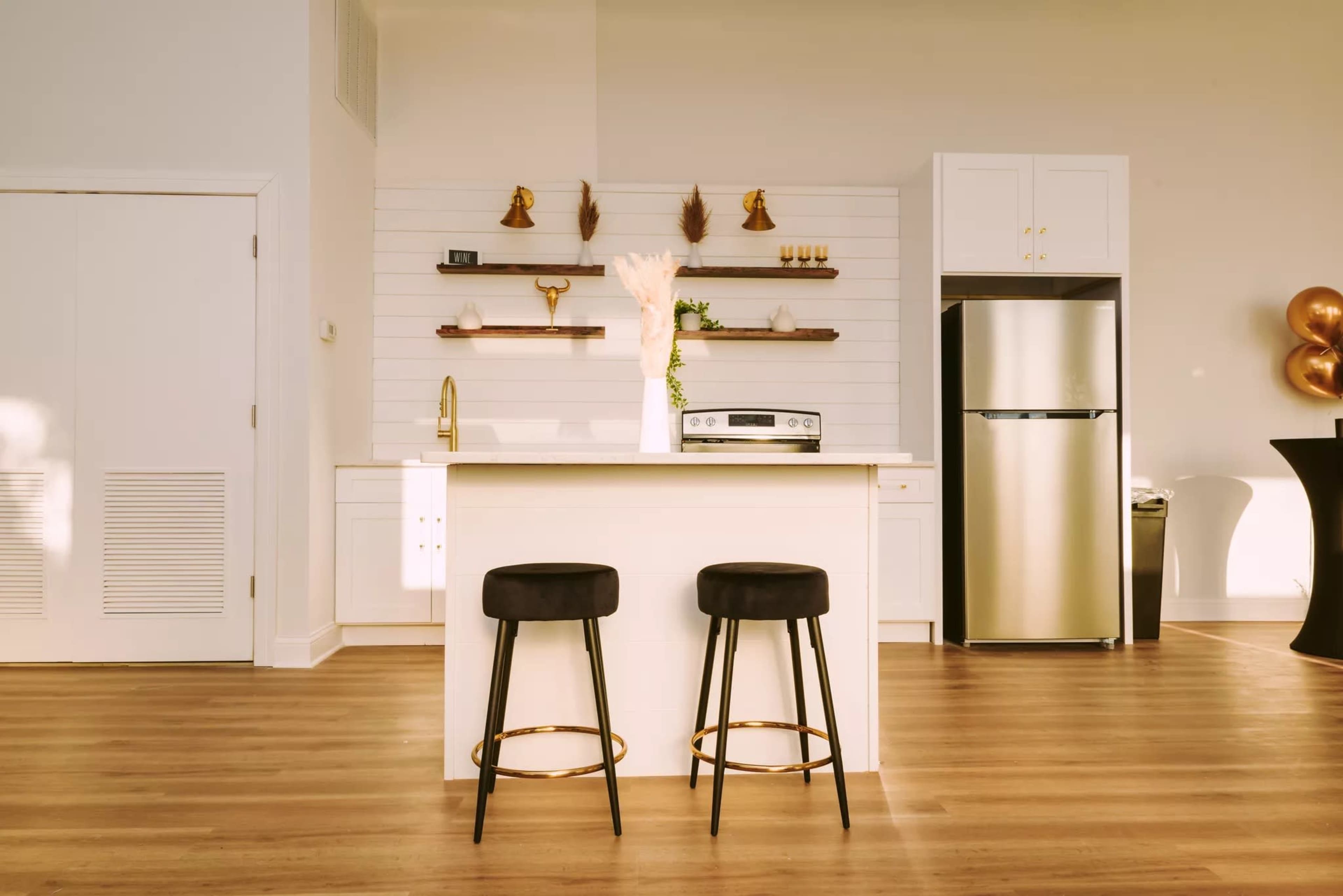 A modern kitchen features a white shiplap wall with floating shelves, an island with two black bar stools, stainless steel appliances, and a minimalist decor.