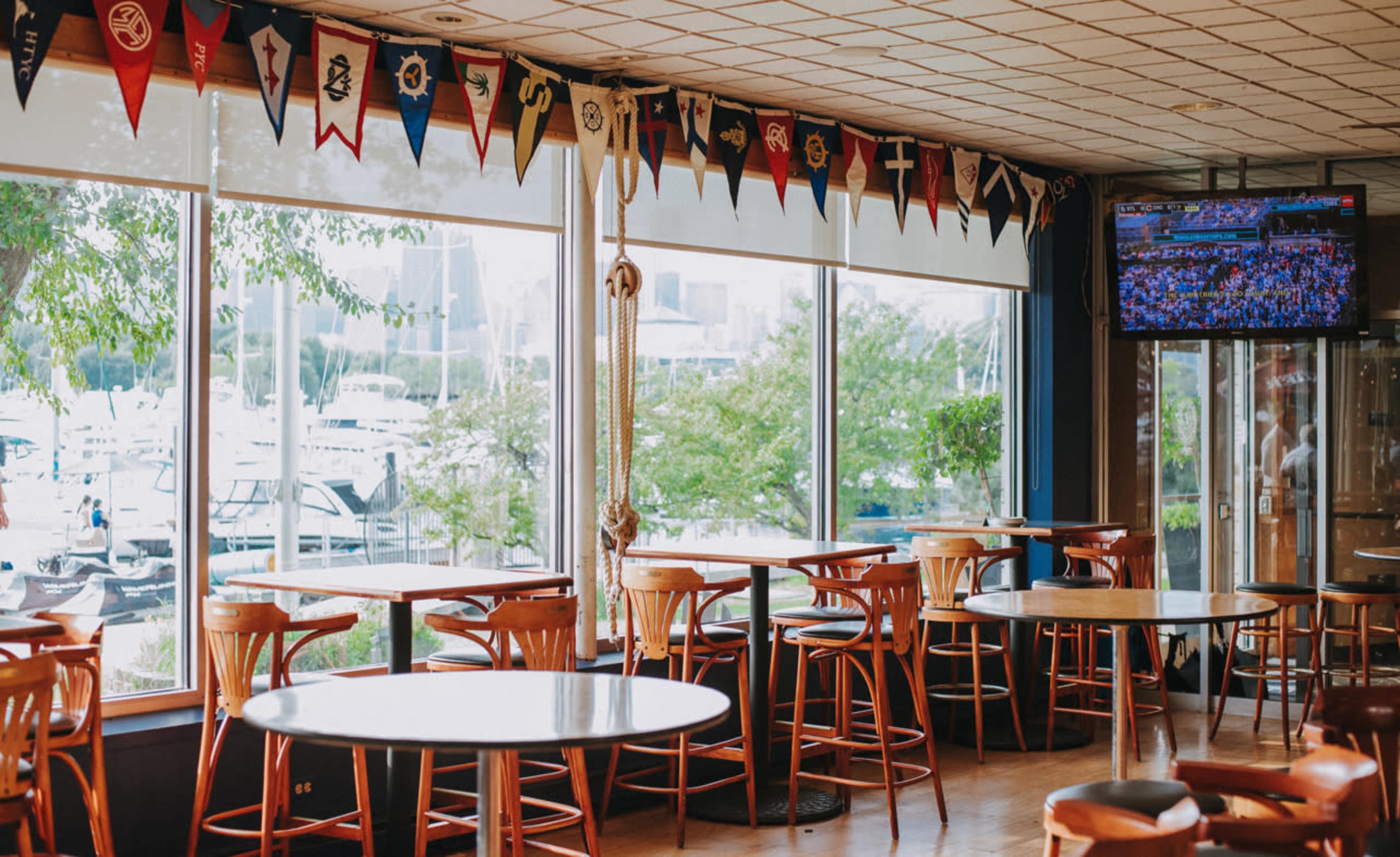 The image shows a waterfront bar with wooden tables and chairs, large windows displaying a marina, and a television showing a sports event.