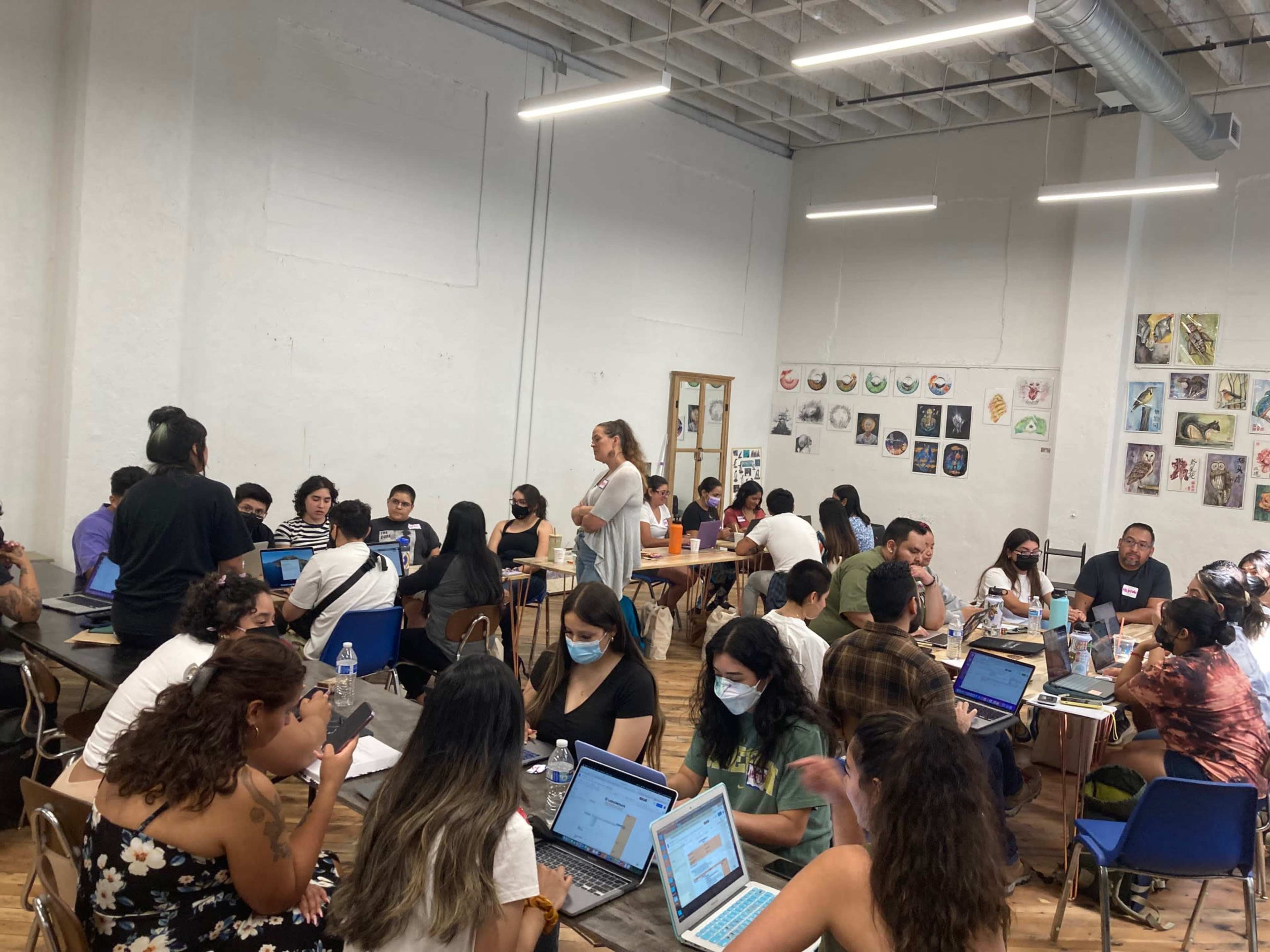 A diverse group of people is engaged in collaborative work at tables in a well-lit room with various artworks displayed on the walls.