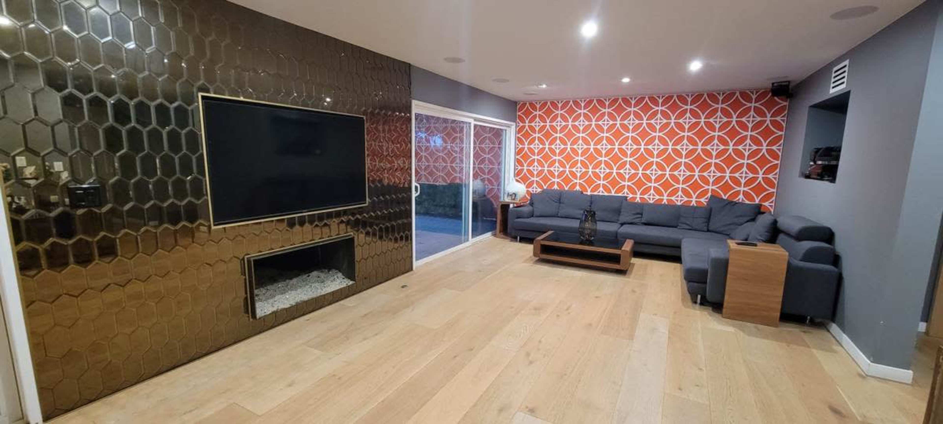 The image depicts a living room with a large television mounted on a hexagonal gold accent wall, a modern gray couch, and a contrasting orange patterned feature wall.