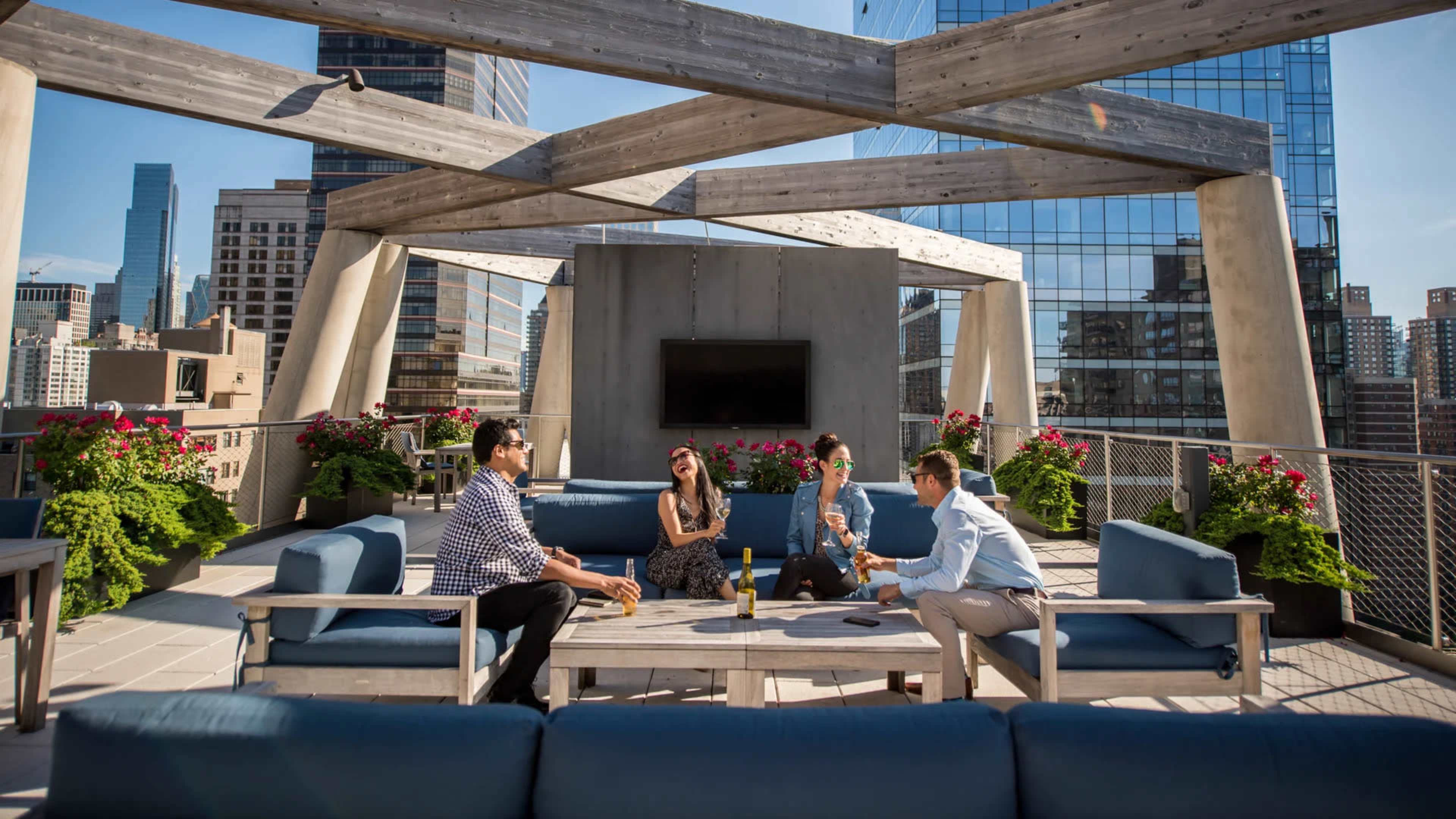 A group of four people is seated around a table on a rooftop terrace surrounded by modern buildings and planters with flowers.