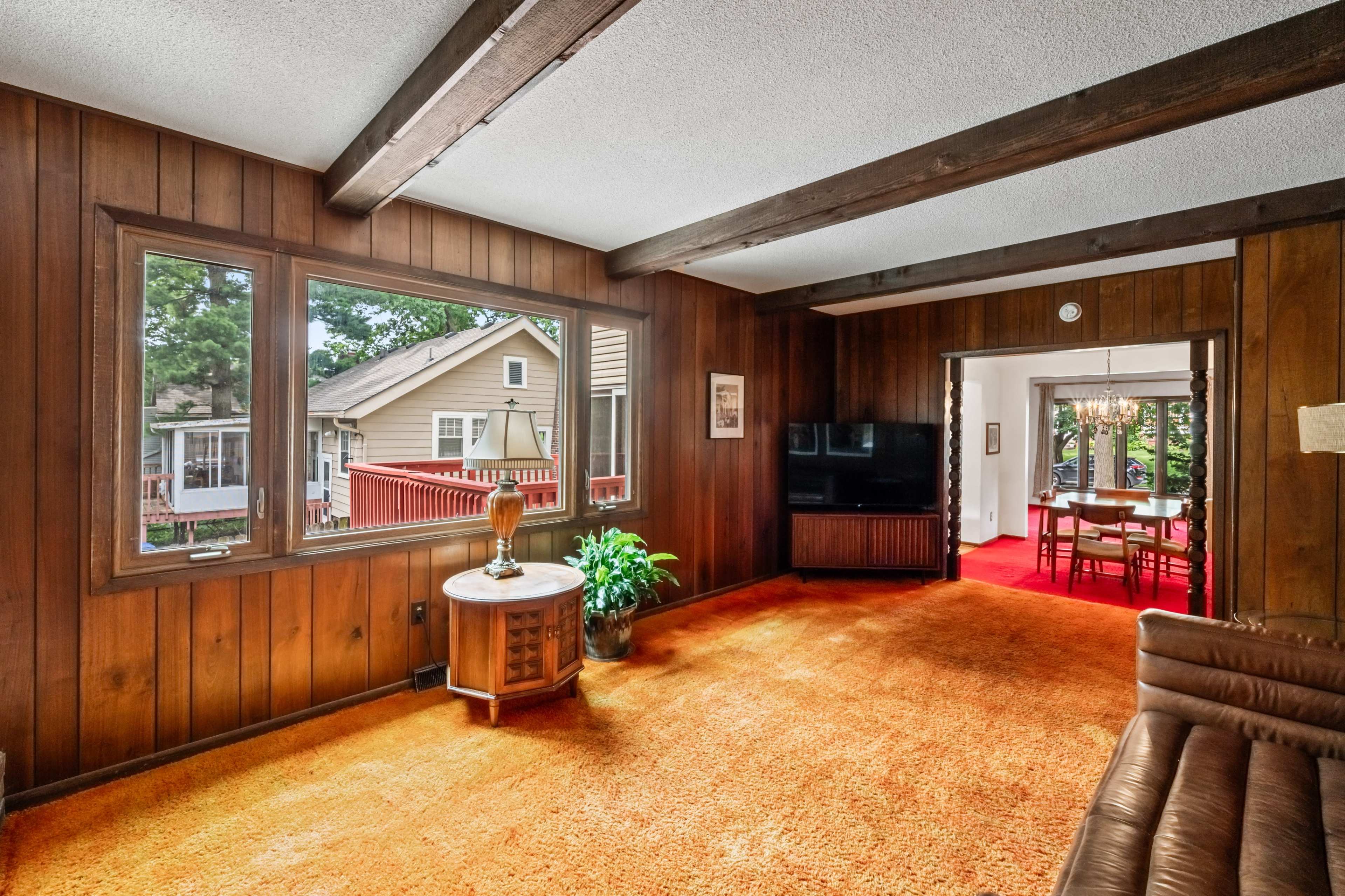 A living room with wooden paneling, large windows, and an adjacent dining area visible through an open doorway.