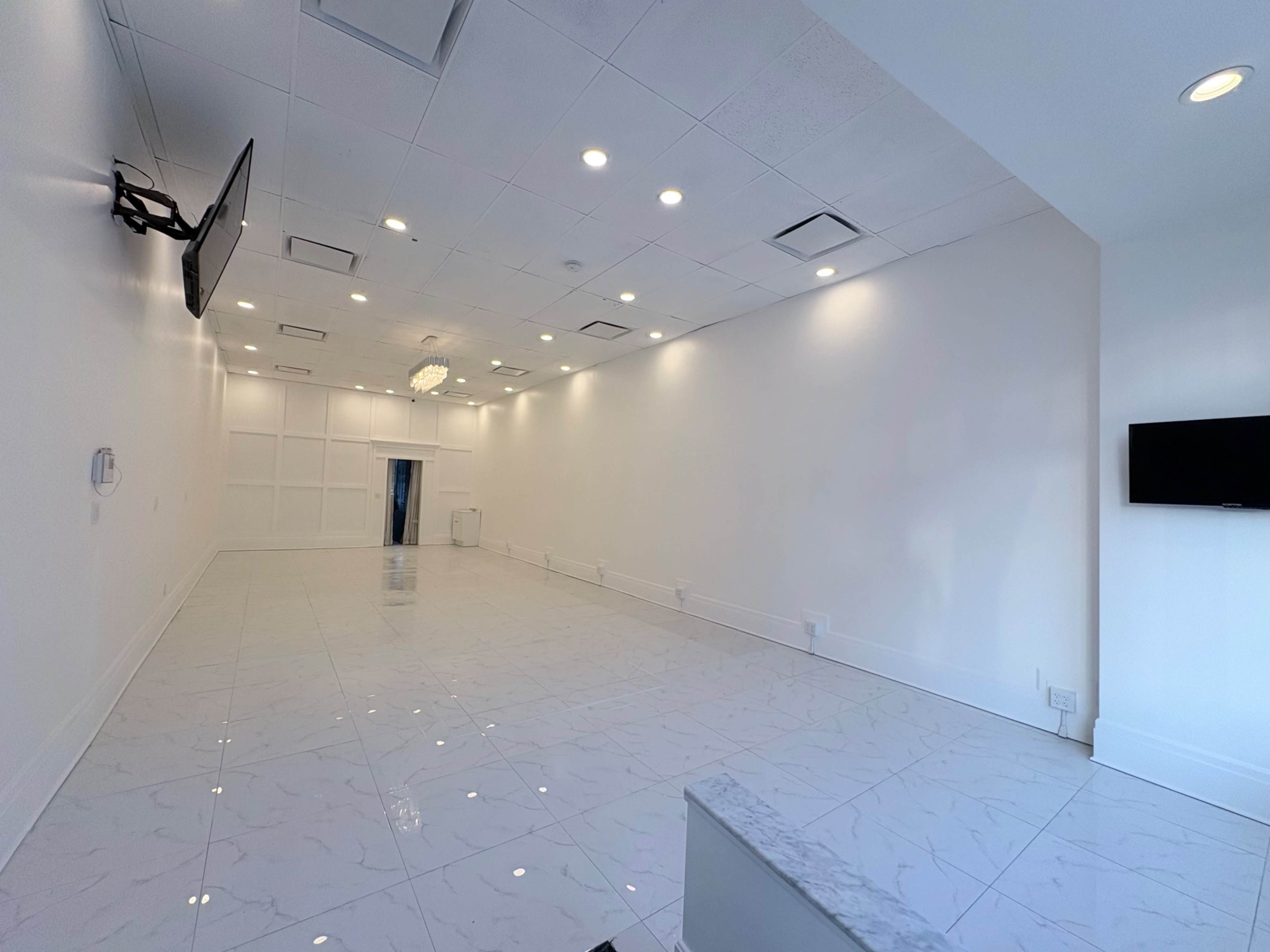 The image shows a spacious, empty room with white walls and glossy tiled flooring, equipped with two mounted televisions.