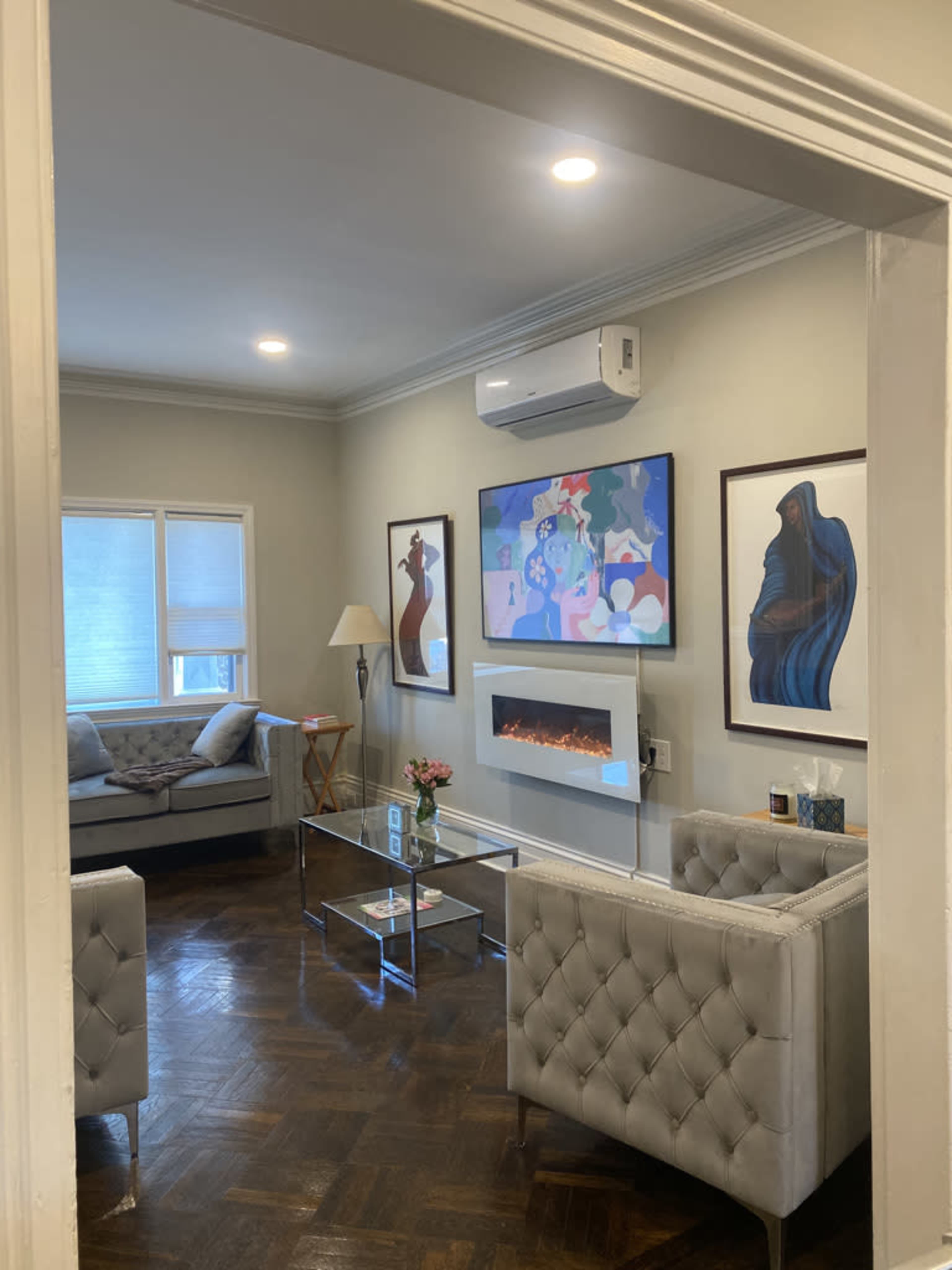 A modern living room with a glass coffee table, two tufted armchairs, a couch, and artwork on the walls, including a contemporary fireplace.