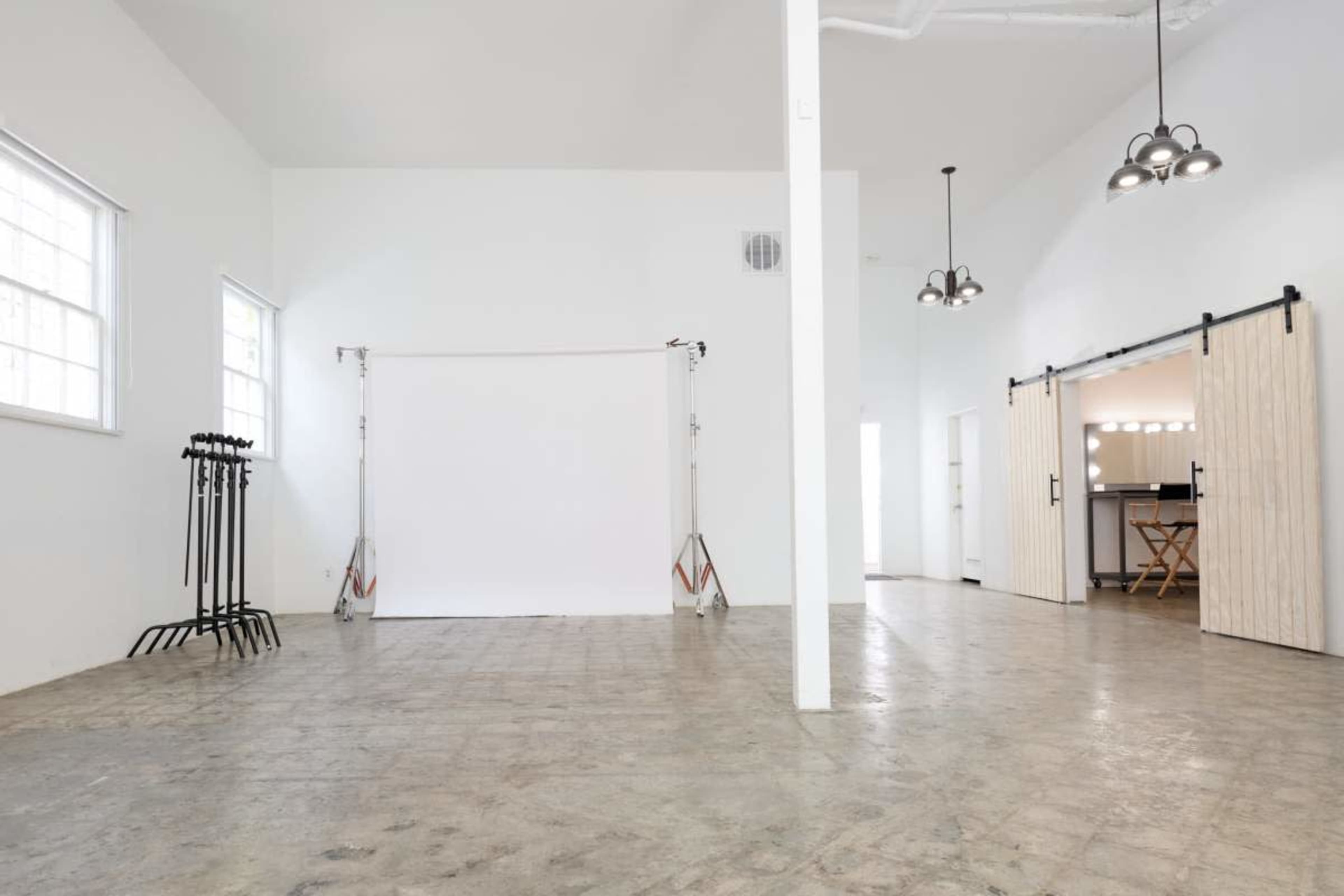 The image shows a spacious, empty studio with a white backdrop, concrete flooring, and large windows, featuring overhead light fixtures and a kitchenette in the background.