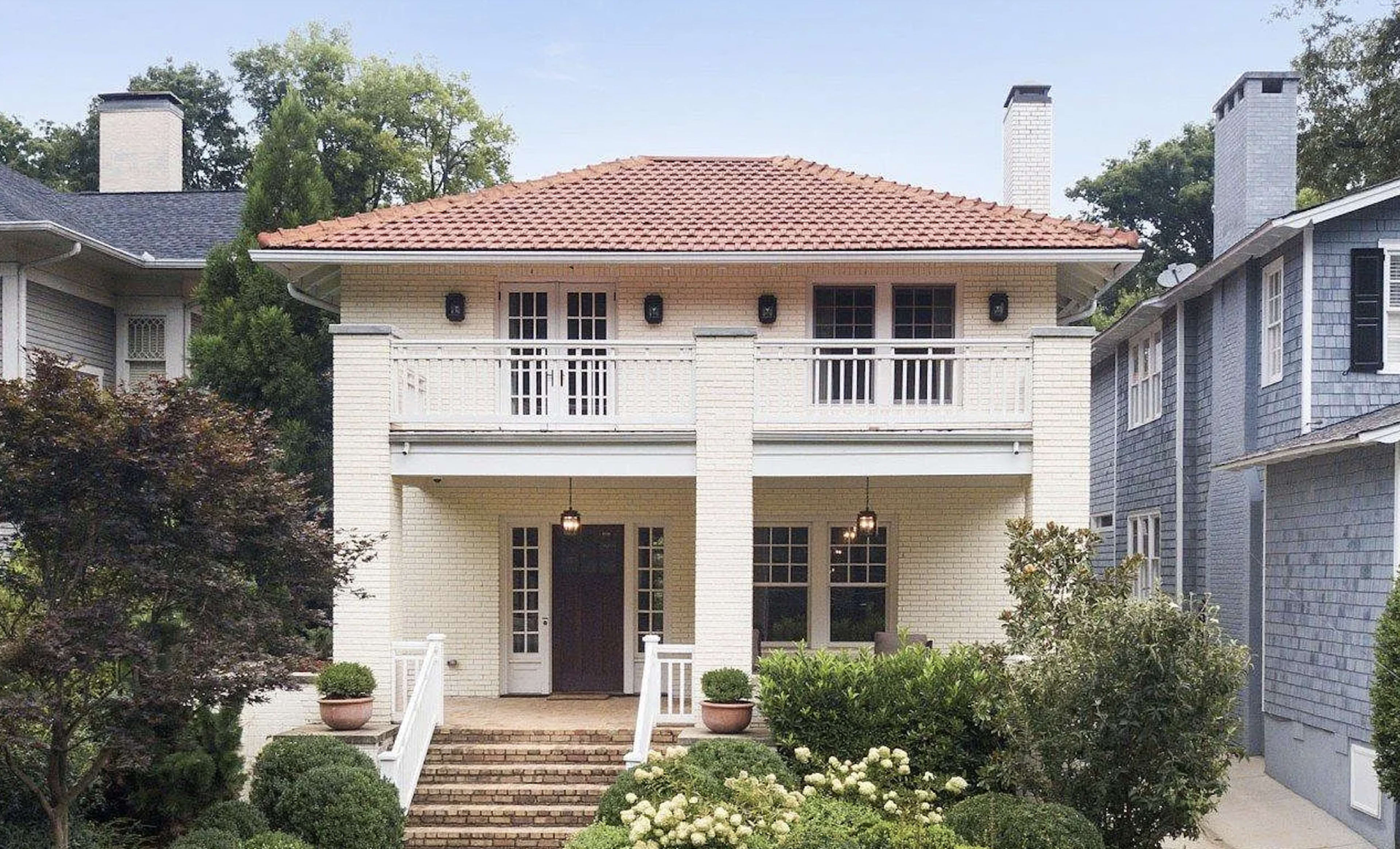A two-story house with a red tiled roof features white brick exterior and a front porch with potted plants on either side of the entrance.