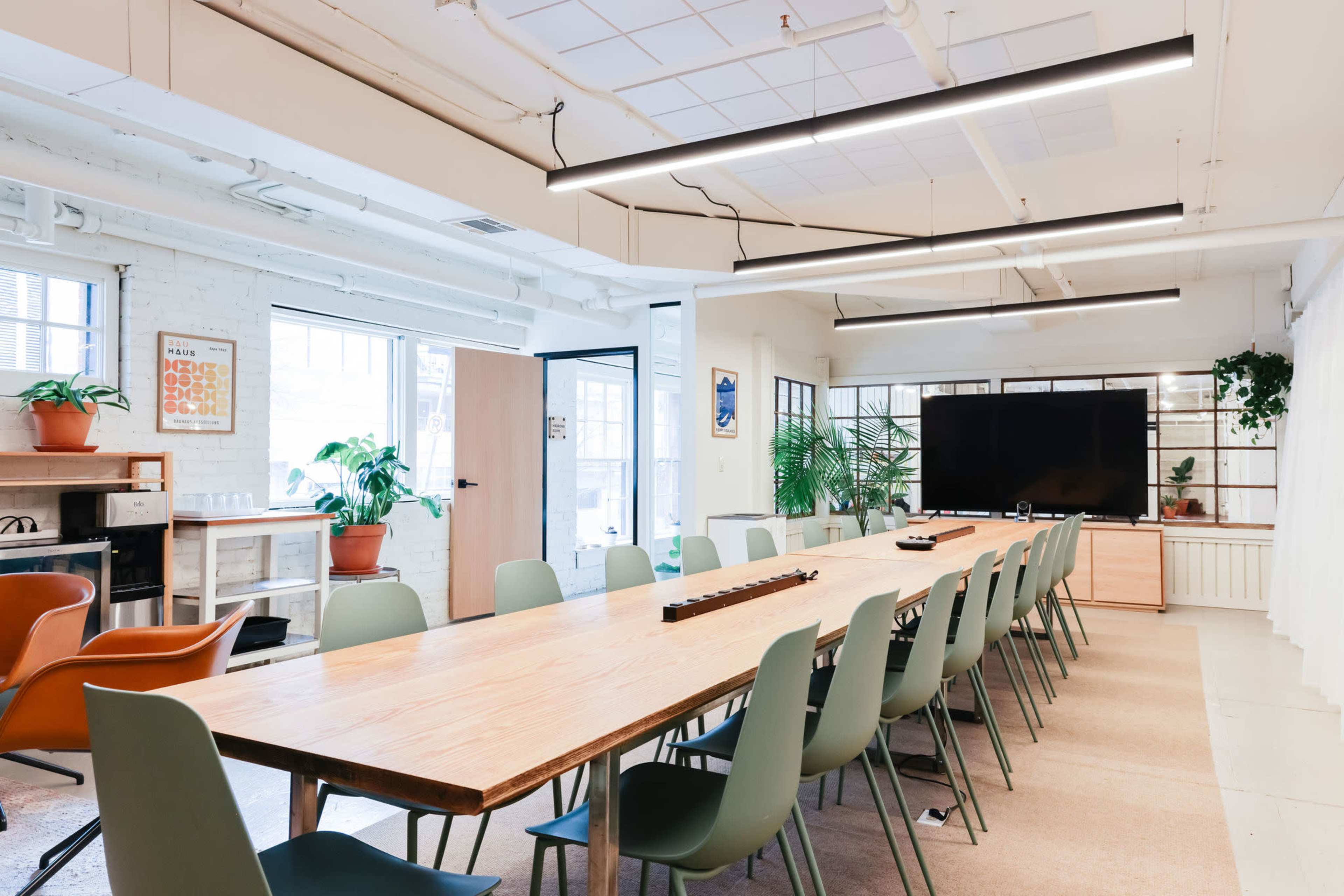 A long wooden table surrounded by green chairs is set up in a bright conference room featuring large windows and a television screen on a wooden stand.