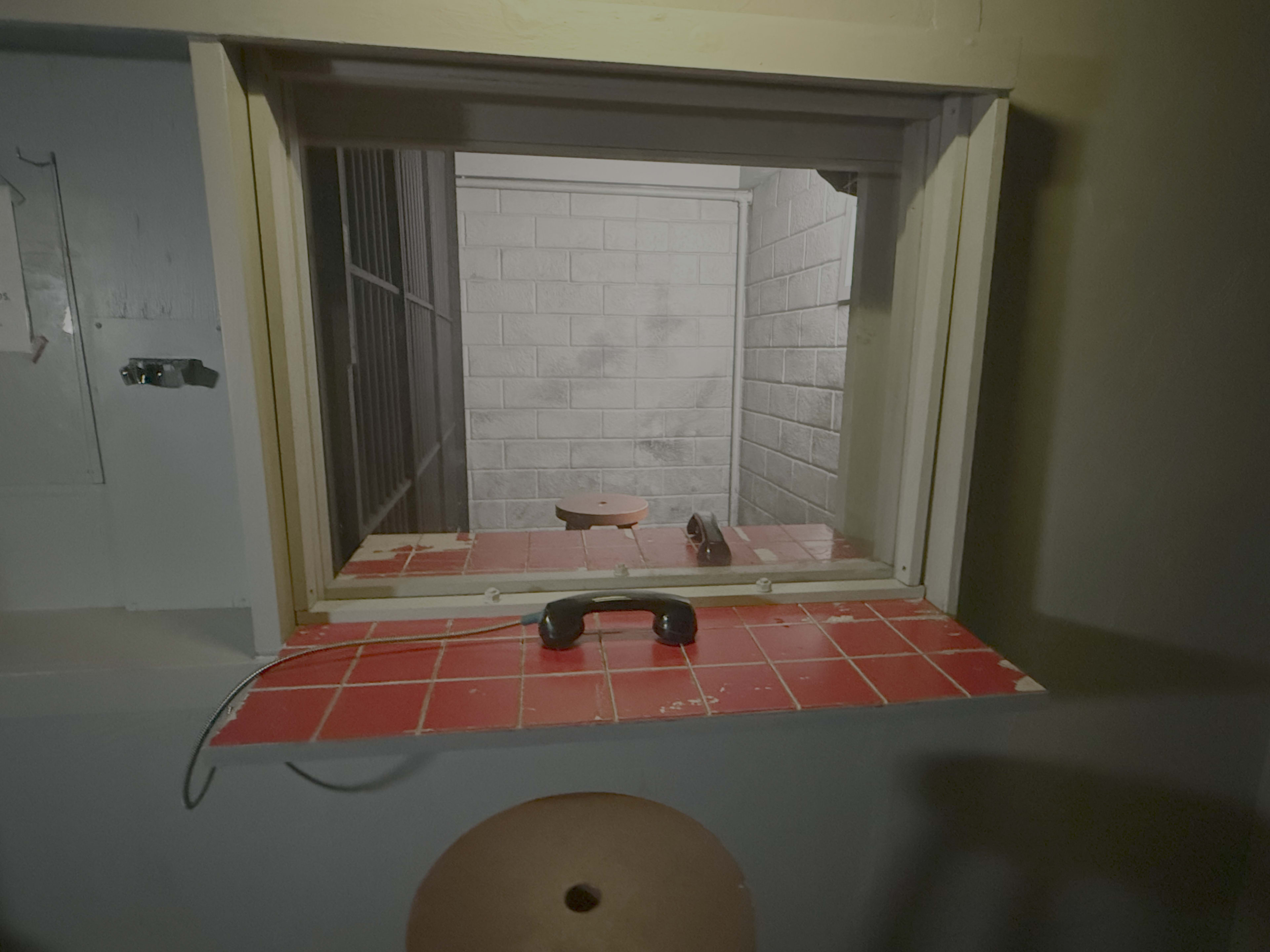 The image shows a view through a window into a small, empty room with concrete walls, featuring a single table and a telephone on a tiled counter.