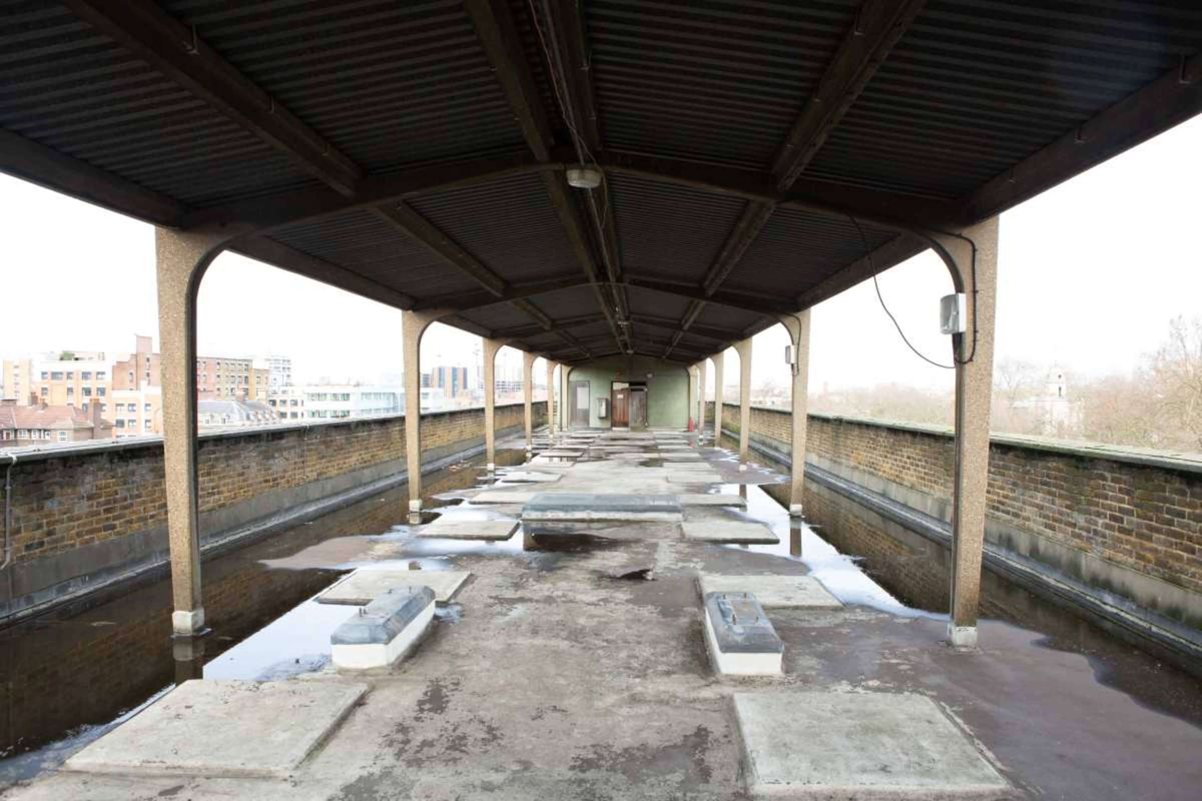 The image shows a long, abandoned corridor on a rooftop with concrete slabs and puddles of water on the ground.