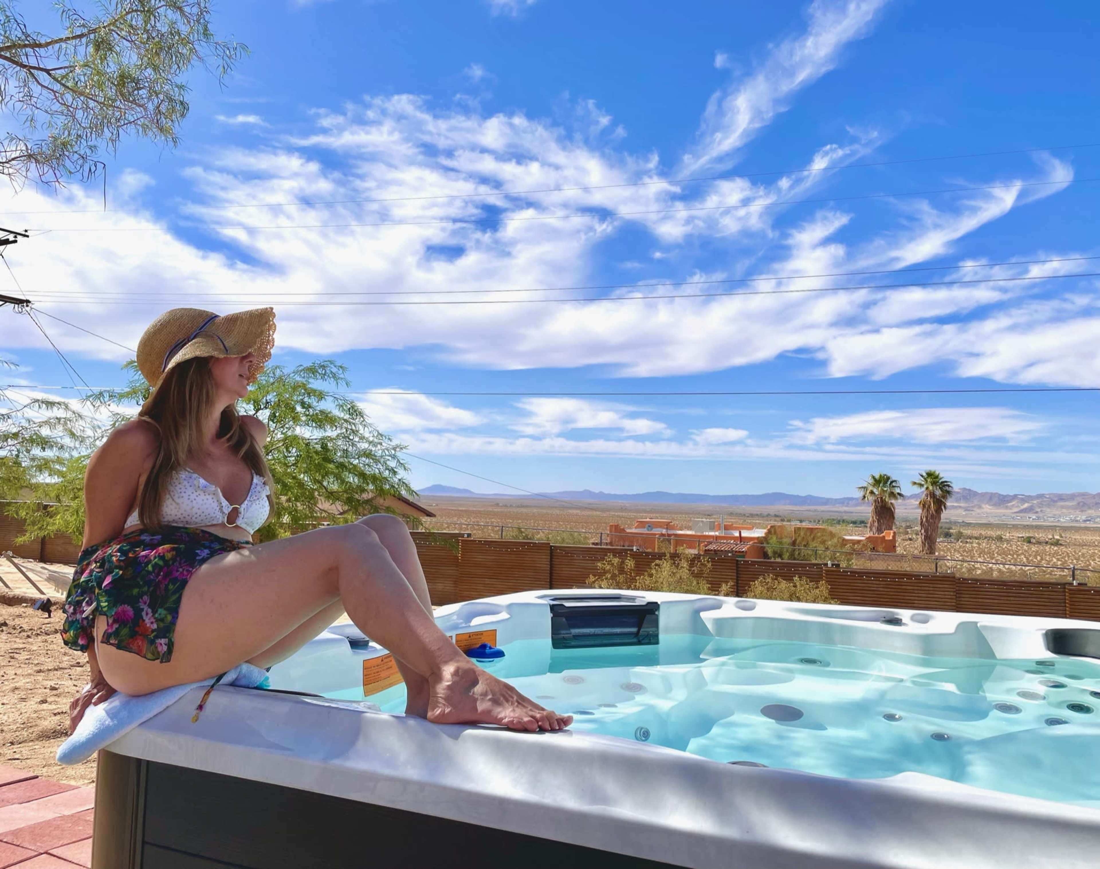 A person in a hat relaxes by a hot tub with a desert landscape and blue sky in the background.