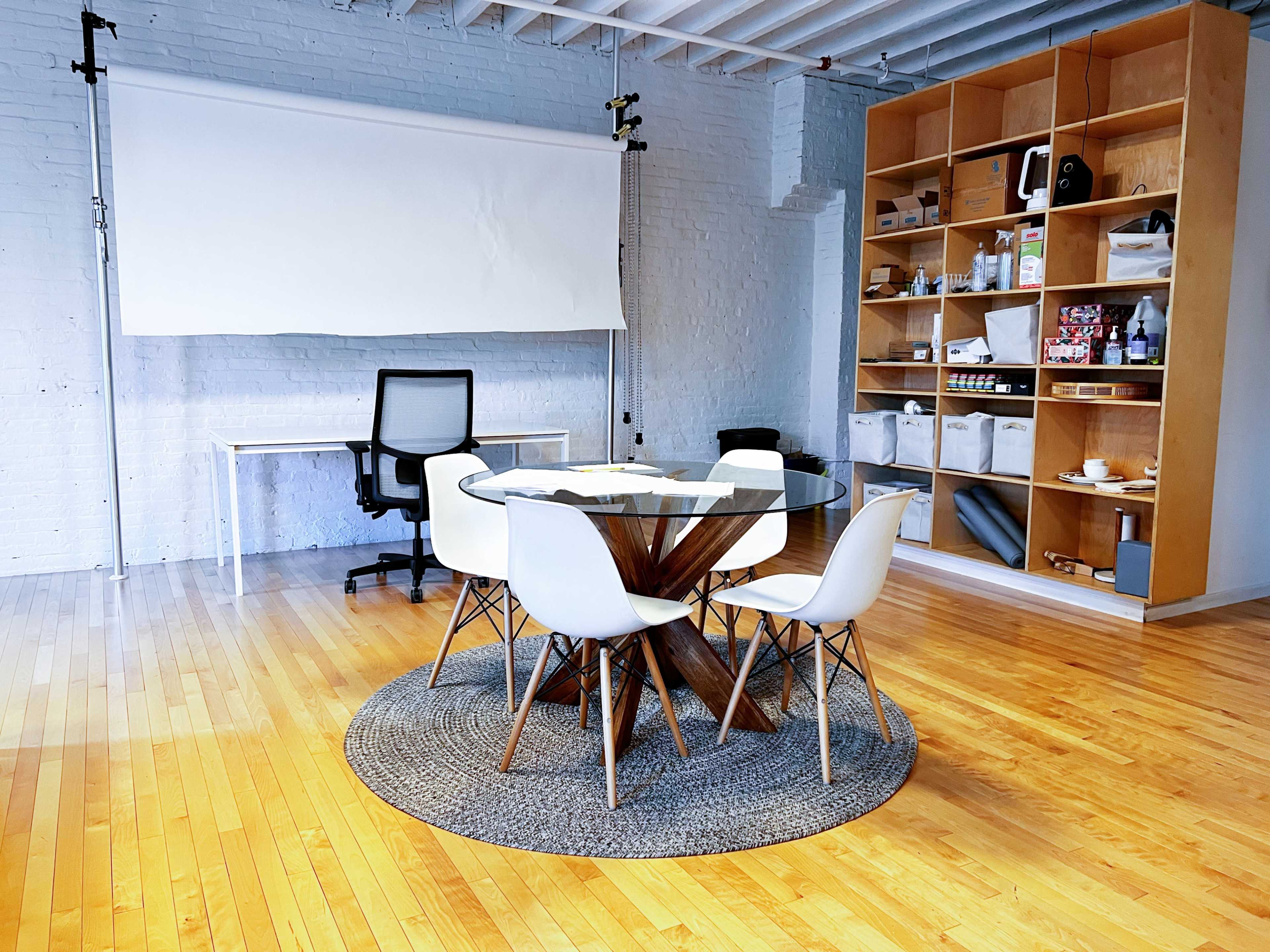 A modern conference room with a round table surrounded by six white chairs, a large wall shelf filled with various items, and a backdrop setup.