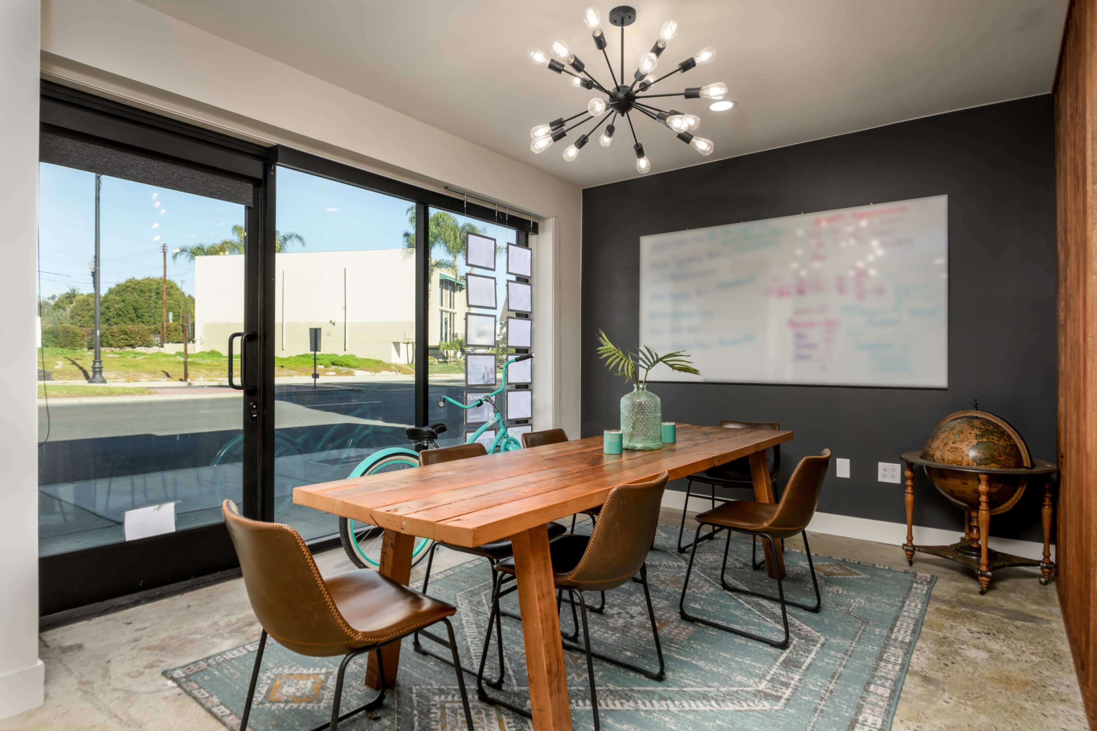 A modern conference room features a wooden table surrounded by leather chairs, large glass doors opening to a view of the street, and a wall-mounted whiteboard.