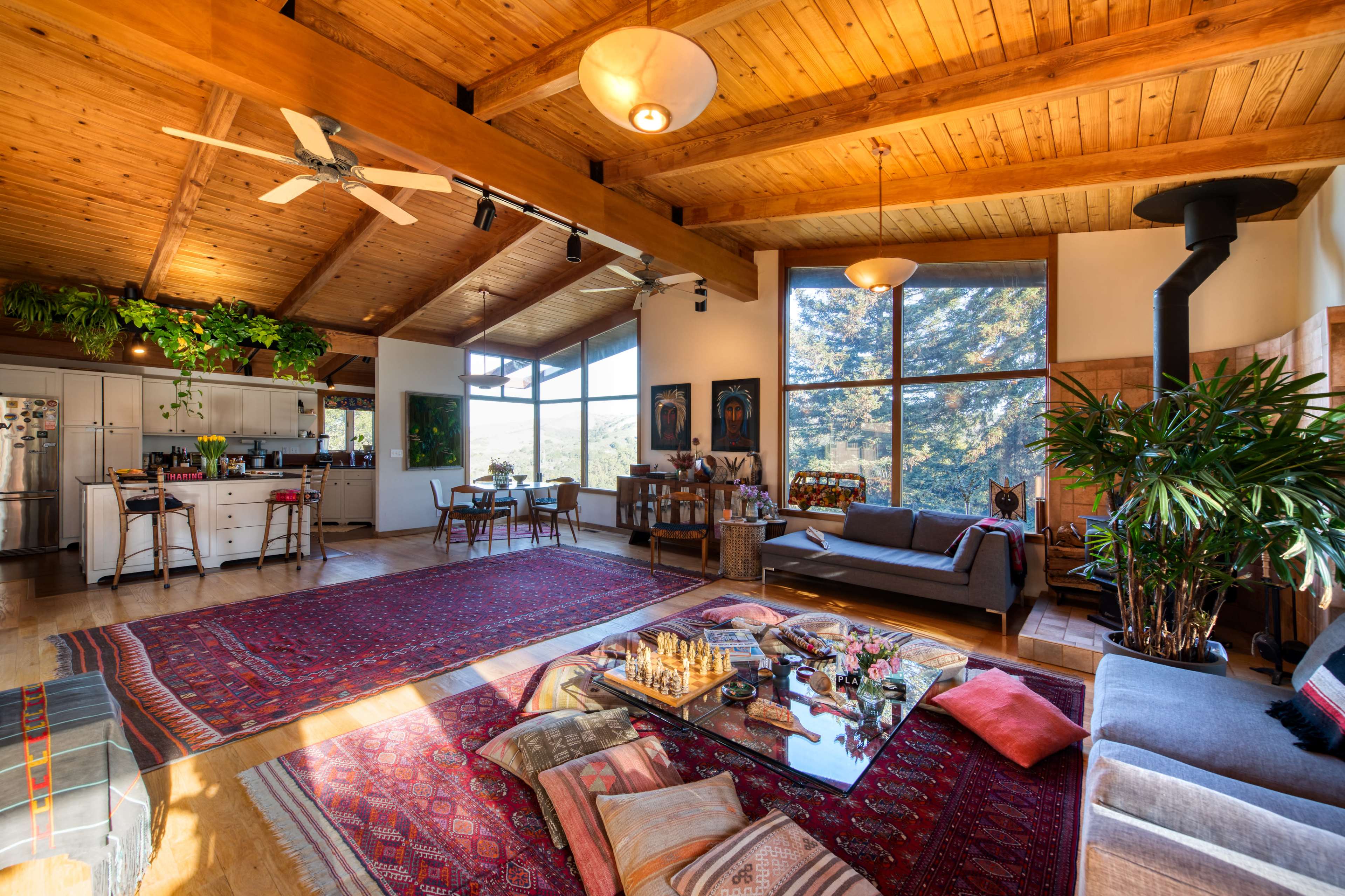 A spacious living area featuring a high wooden ceiling, large windows, a kitchen area, comfortable seating, and various decorative plants.