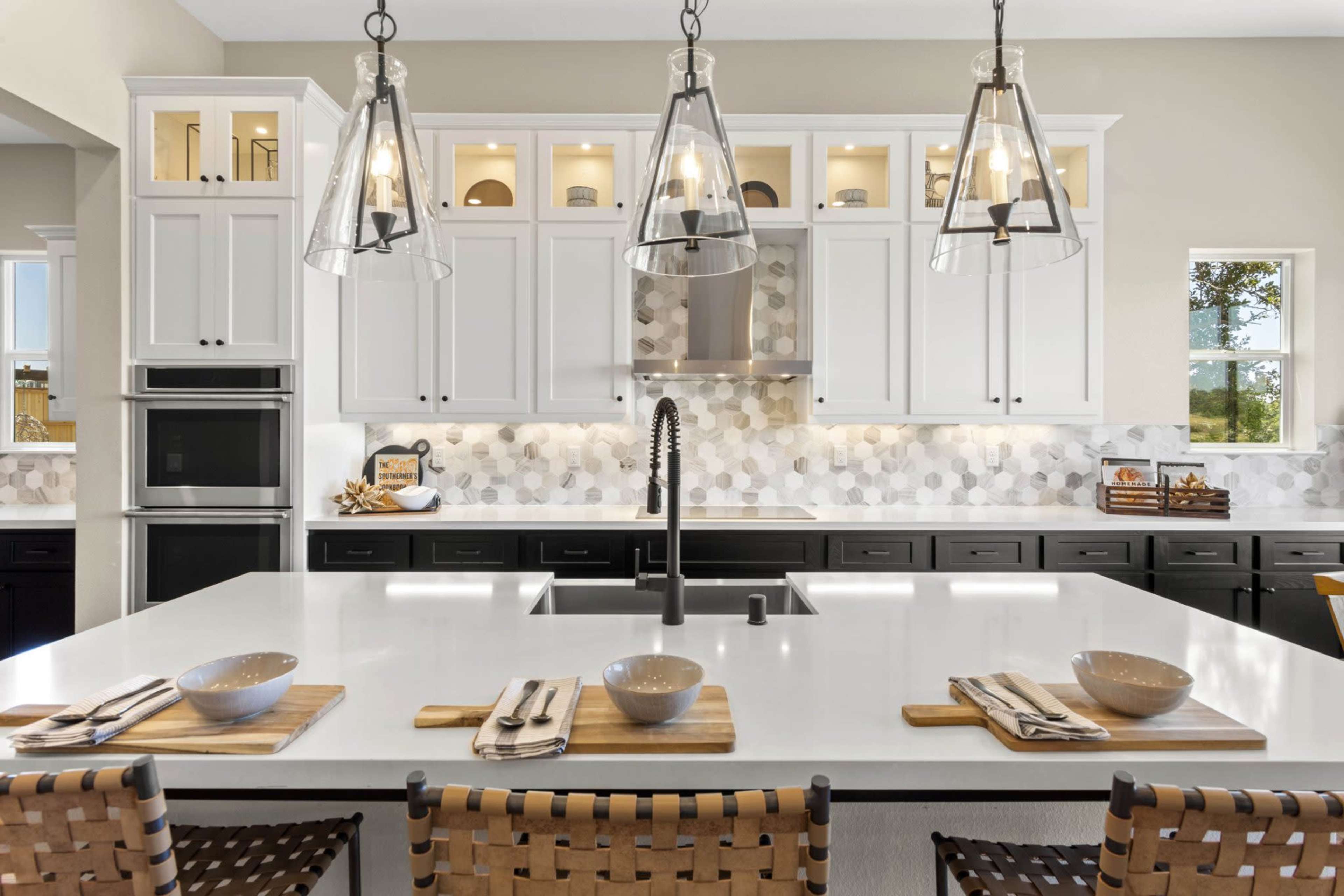 The image shows a modern kitchen with a large island featuring four place settings, white cabinets, a stainless steel hood, and pendant lights.