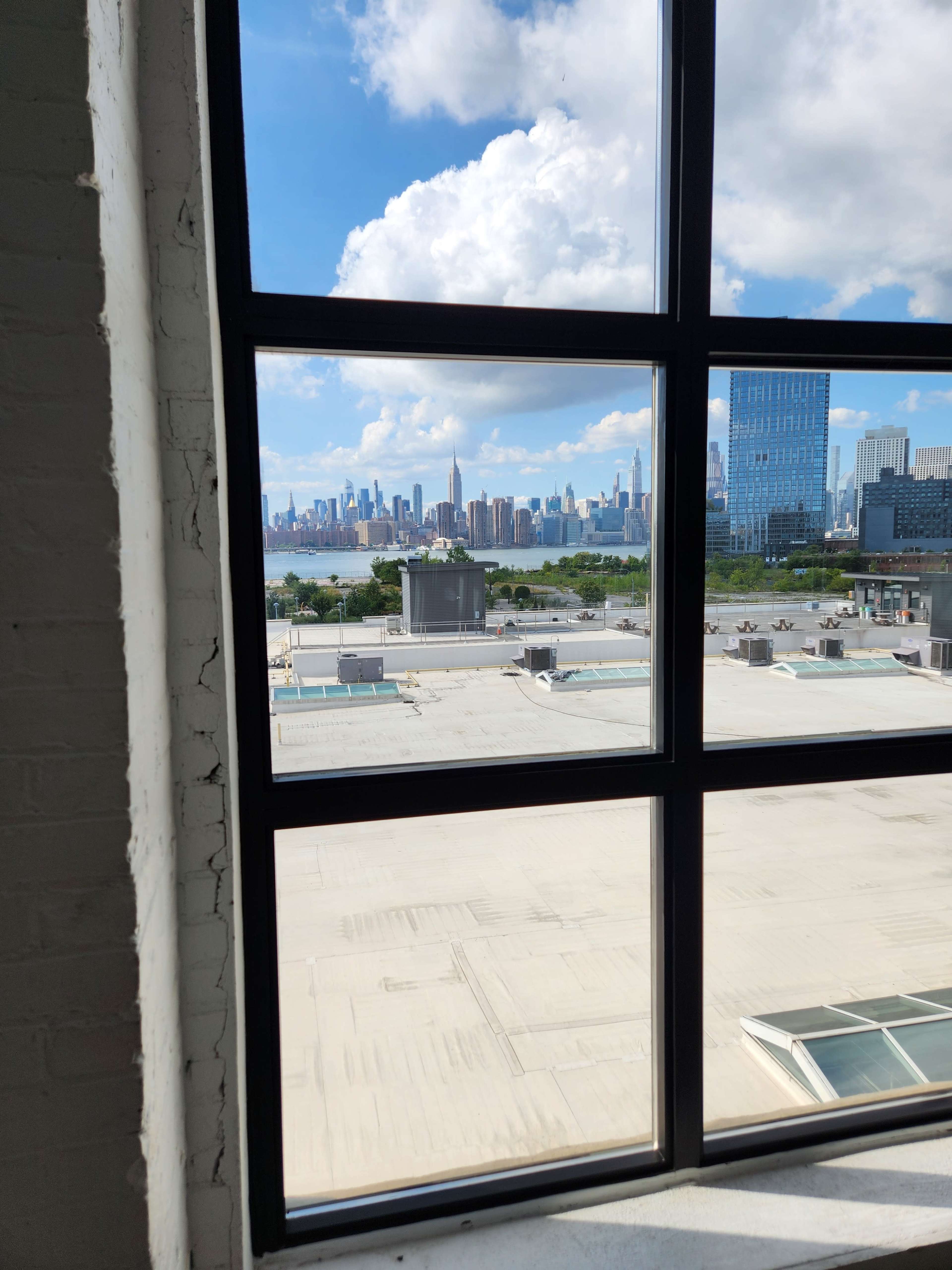 The image shows a large window frame with a view of a city skyline featuring tall buildings and a partly cloudy sky.