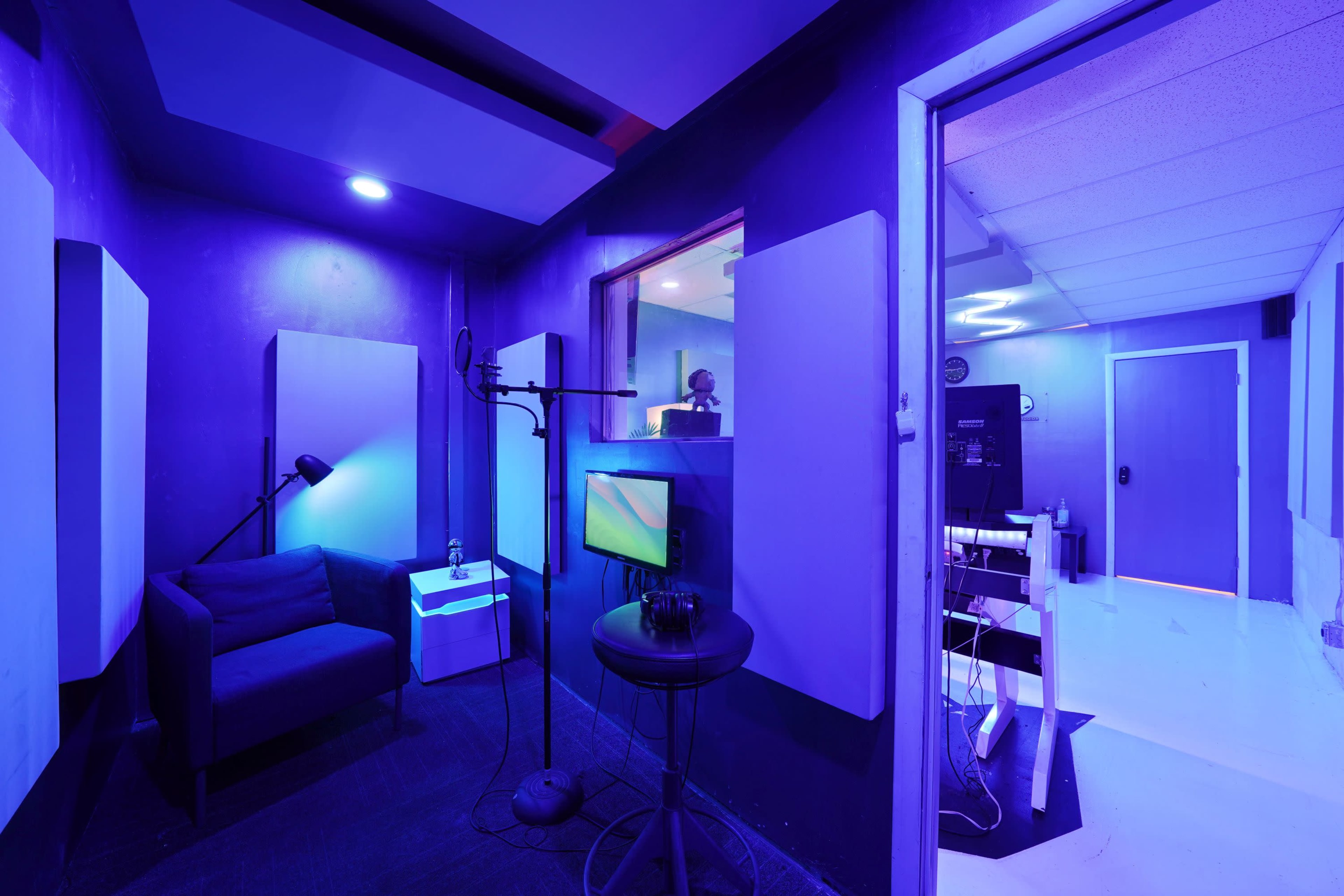 The image depicts a recording studio with purple walls, soundproof panels, a microphone setup, and a visible control room through a glass window.