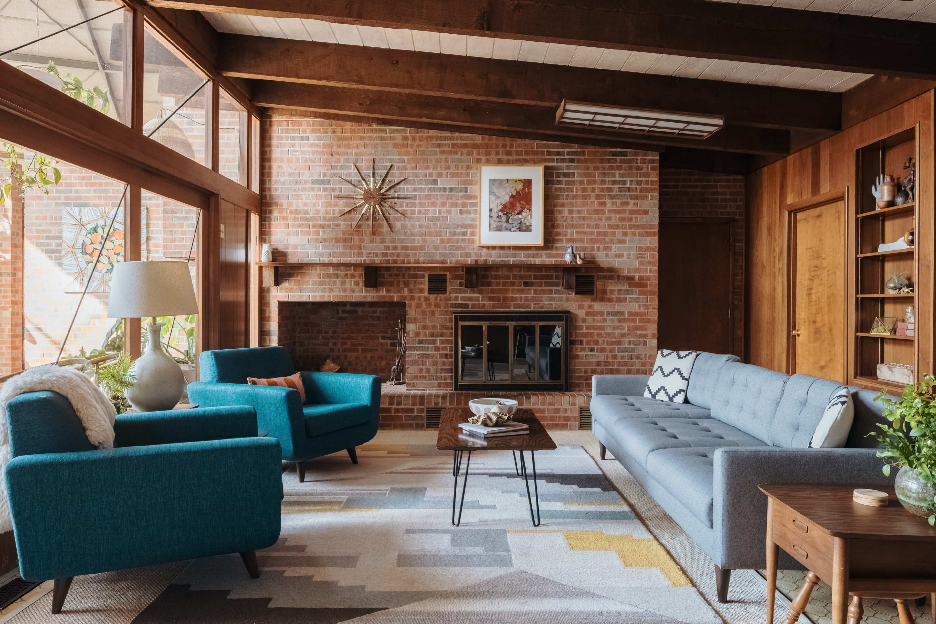 A modern living room with a brick wall, a fireplace, and a mix of teal and gray furniture arranged around a geometric area rug.
