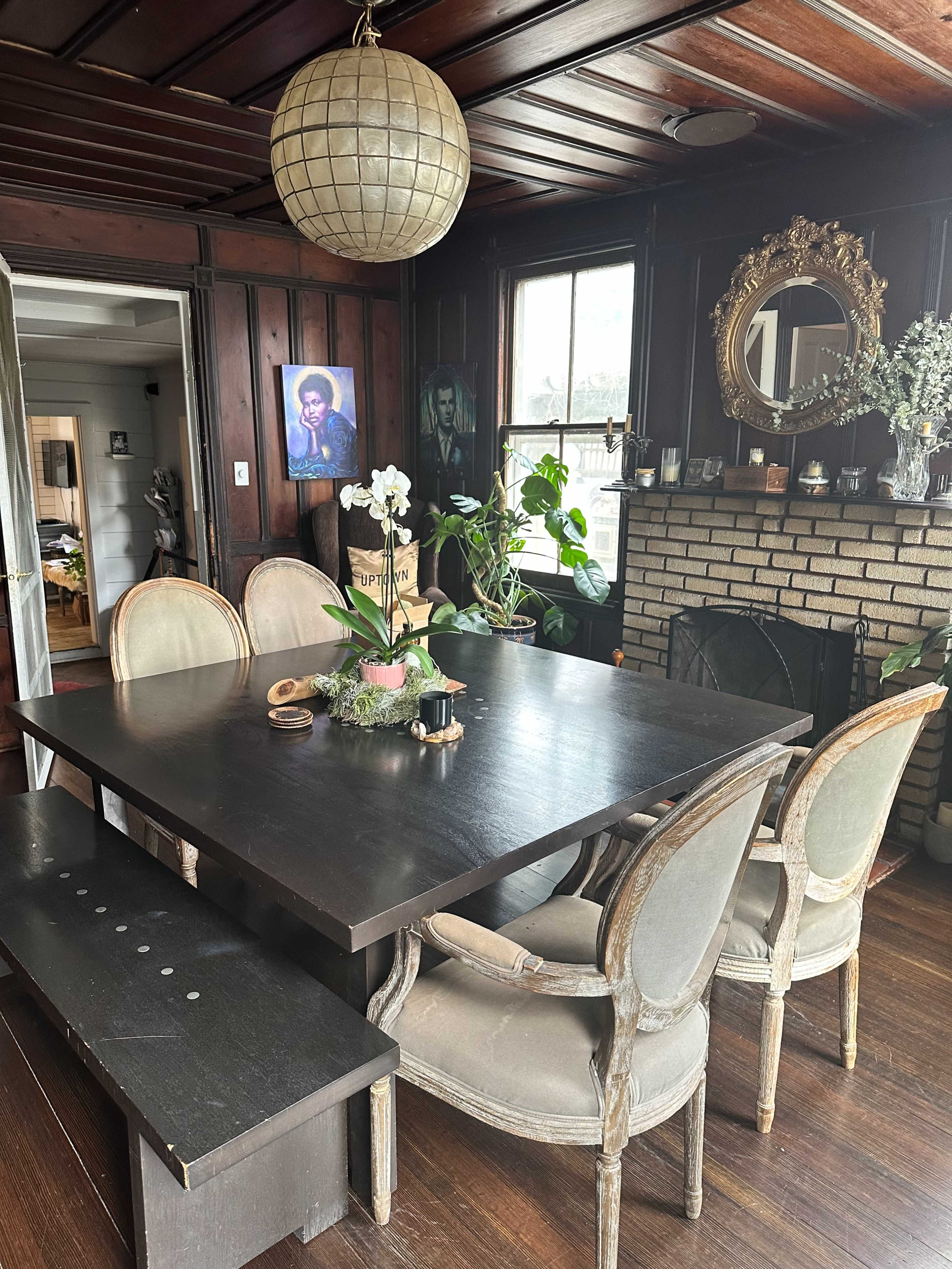A dark wood dining table is surrounded by six chairs, with a large round pendant light hanging above and a decorative mirror on the wall beside a fireplace.