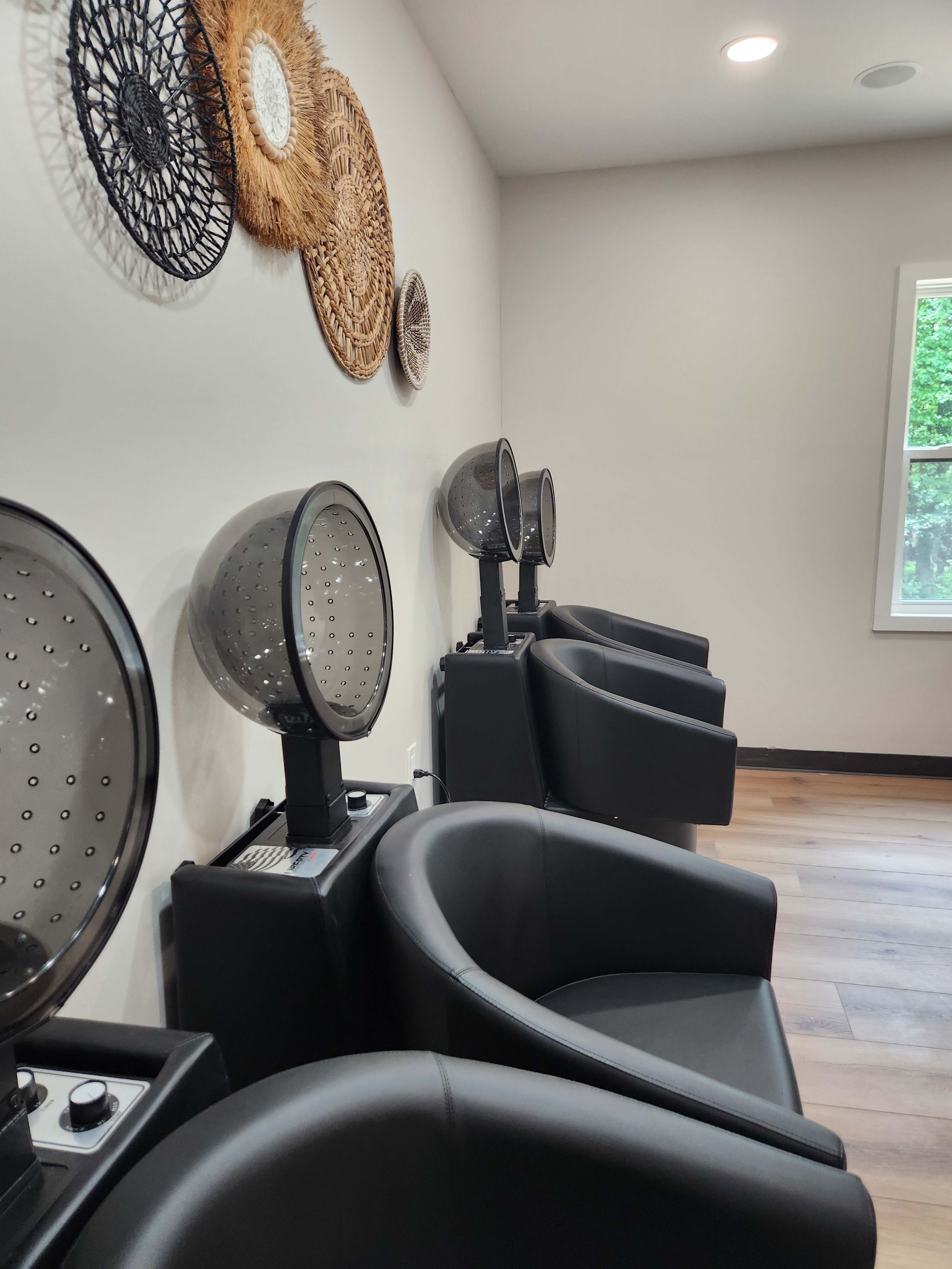 The image shows a salon interior with four black chairs equipped with hooded hair dryers, arranged neatly against a wall adorned with decorative woven wall hangings.