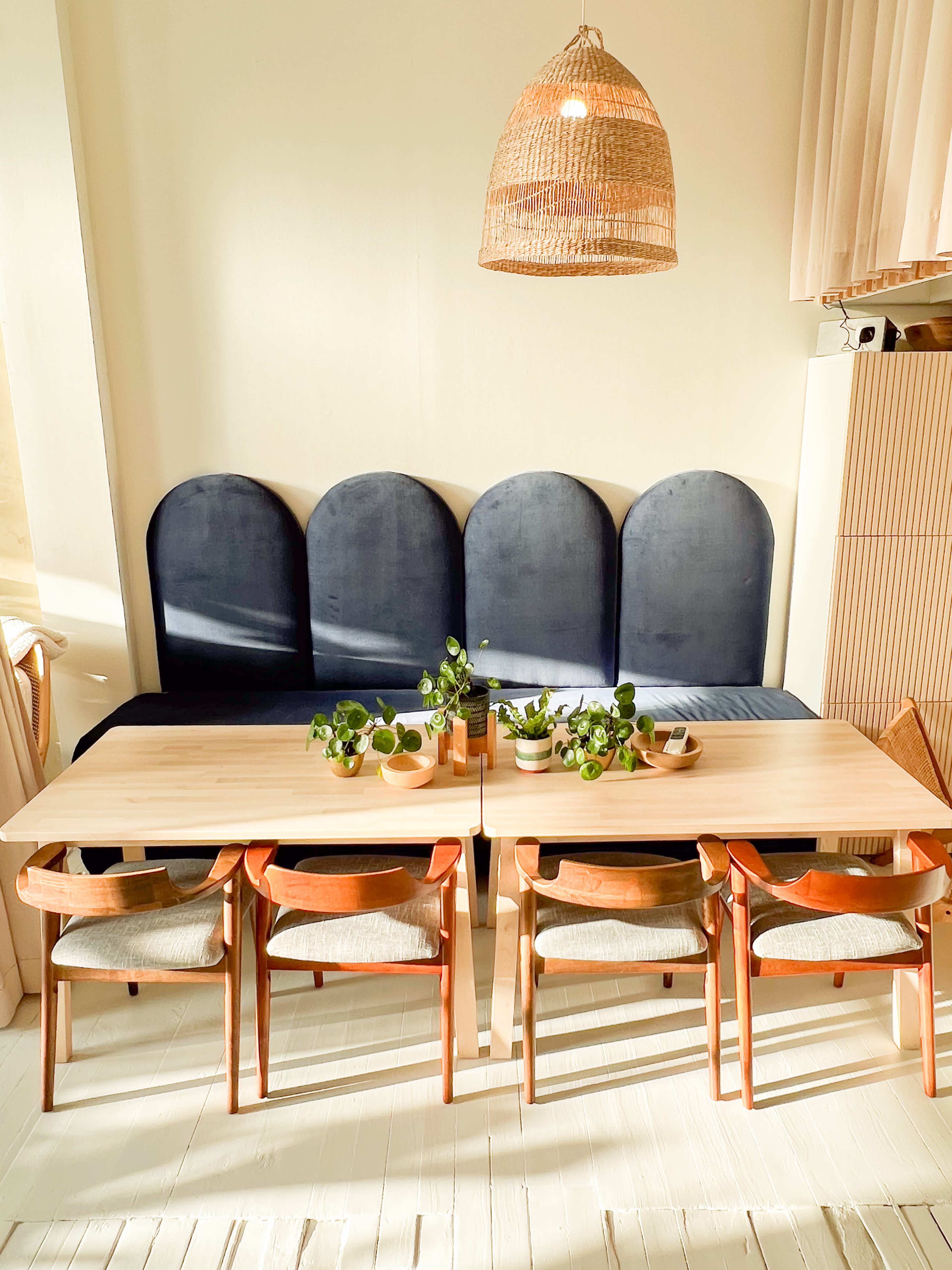 A brightly lit dining area features two wooden tables set with plants, surrounded by chairs facing a cushioned bench along the wall.
