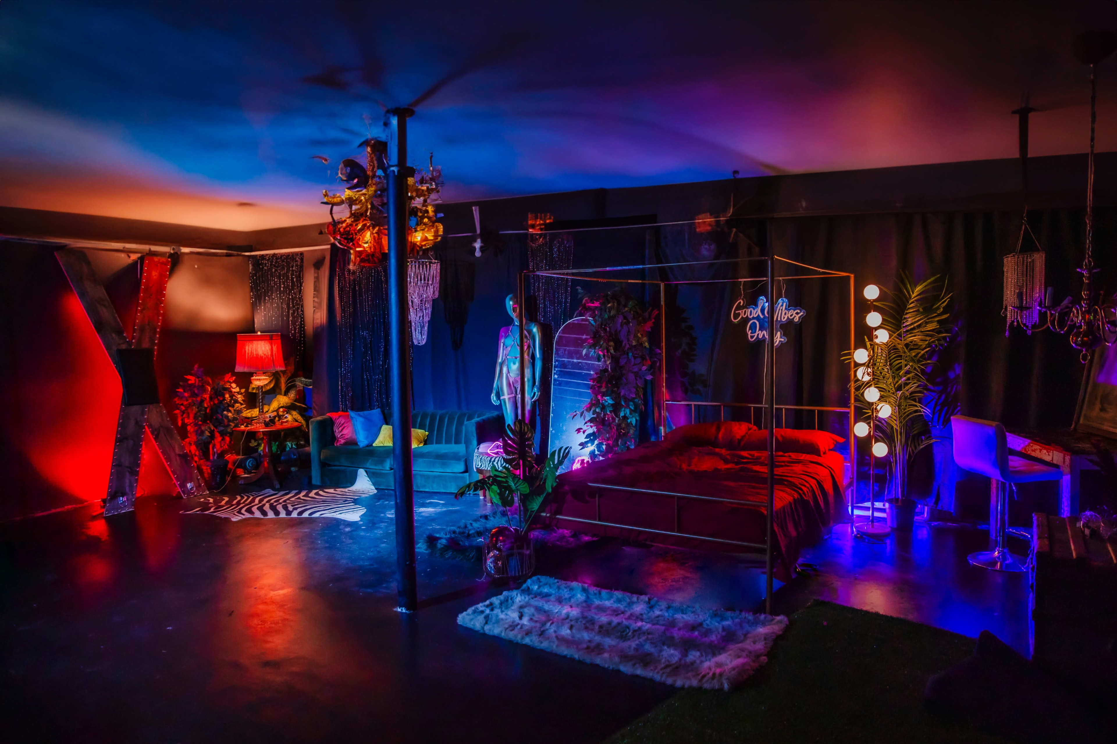 The image shows a dimly lit room with colorful neon lights, a bed with an ornate frame, and various decorations including plants and furniture.