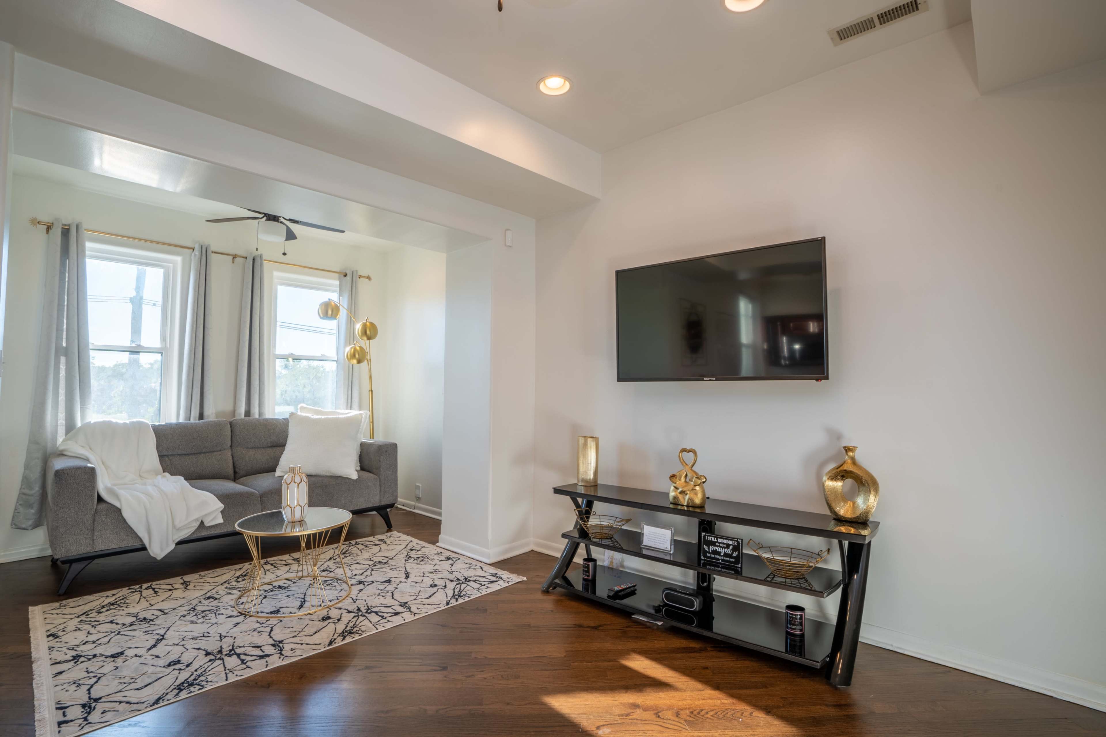 A modern living room features a gray sofa, a black coffee table, and a mounted television against a white wall, with large windows allowing natural light to enter.