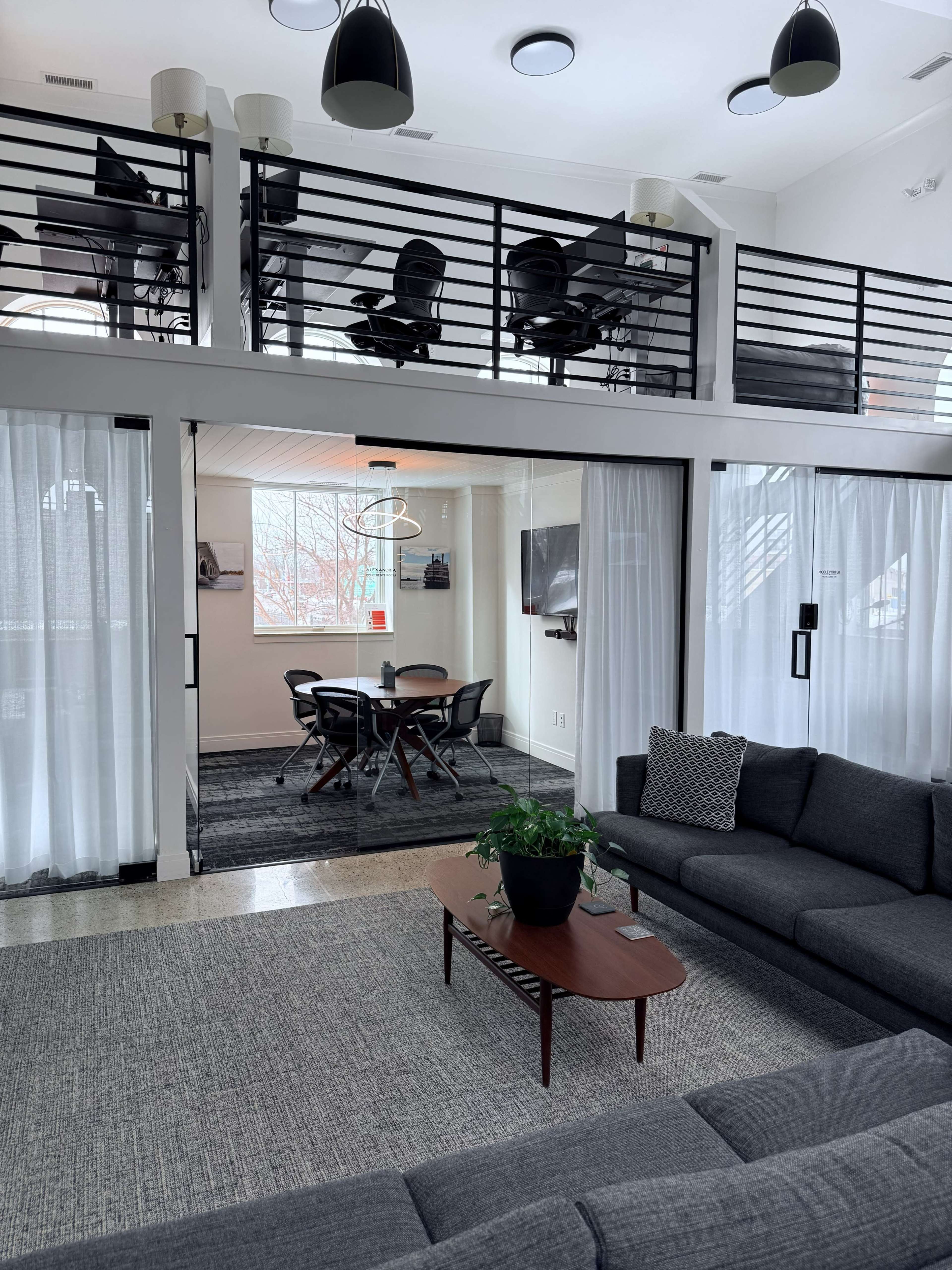 The image shows a modern, open-concept living space with a seating area, a dining table, and a balcony overlooking an office setup.