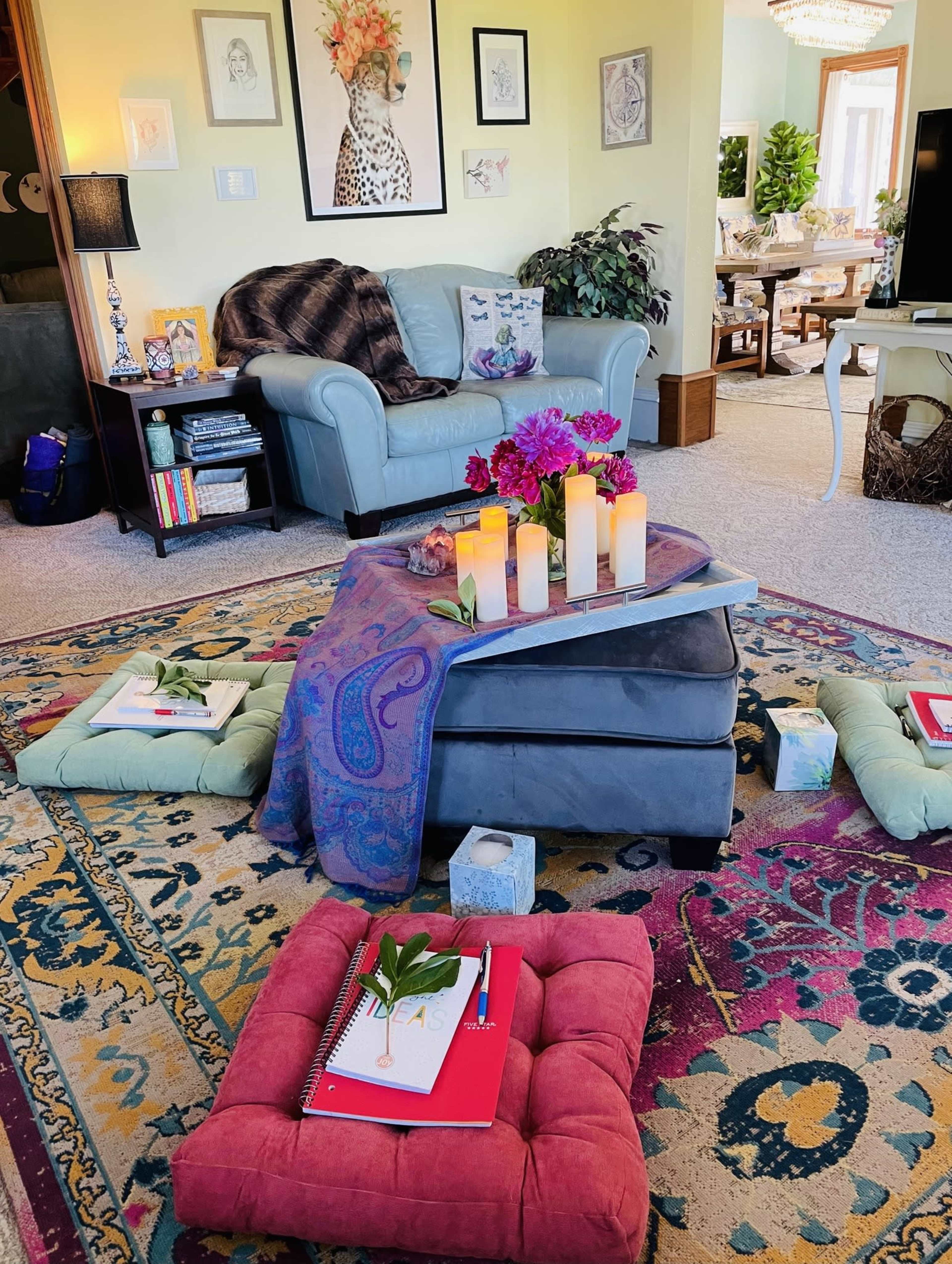 A cozy living room scene features a light blue armchair, a coffee table adorned with candles and flowers, and colorful floor cushions scattered on a patterned rug.
